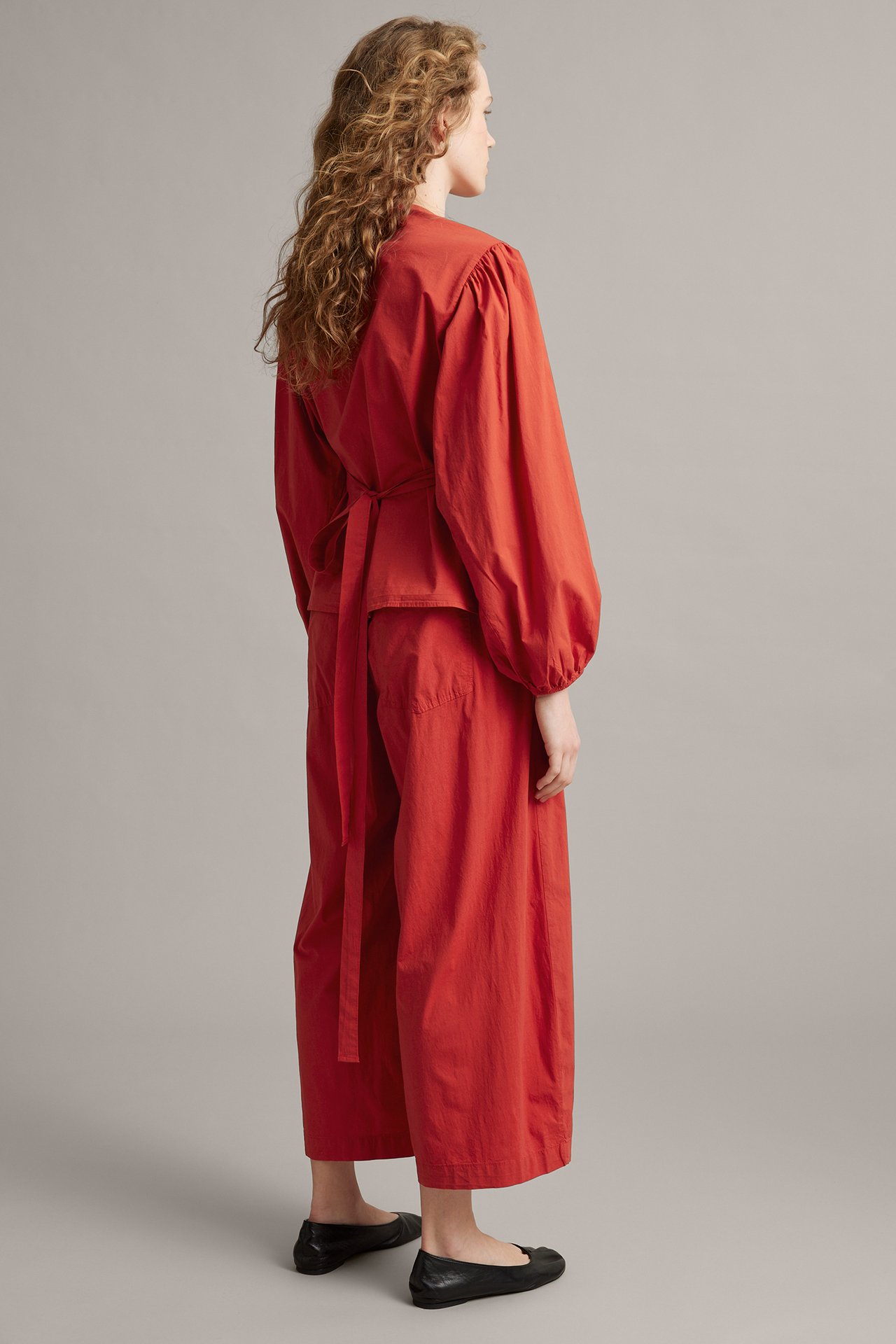 Side-bakfra: kvinne i rød barrel-fit bukse, matchende omslagsbluse, svarte ballerinasko.