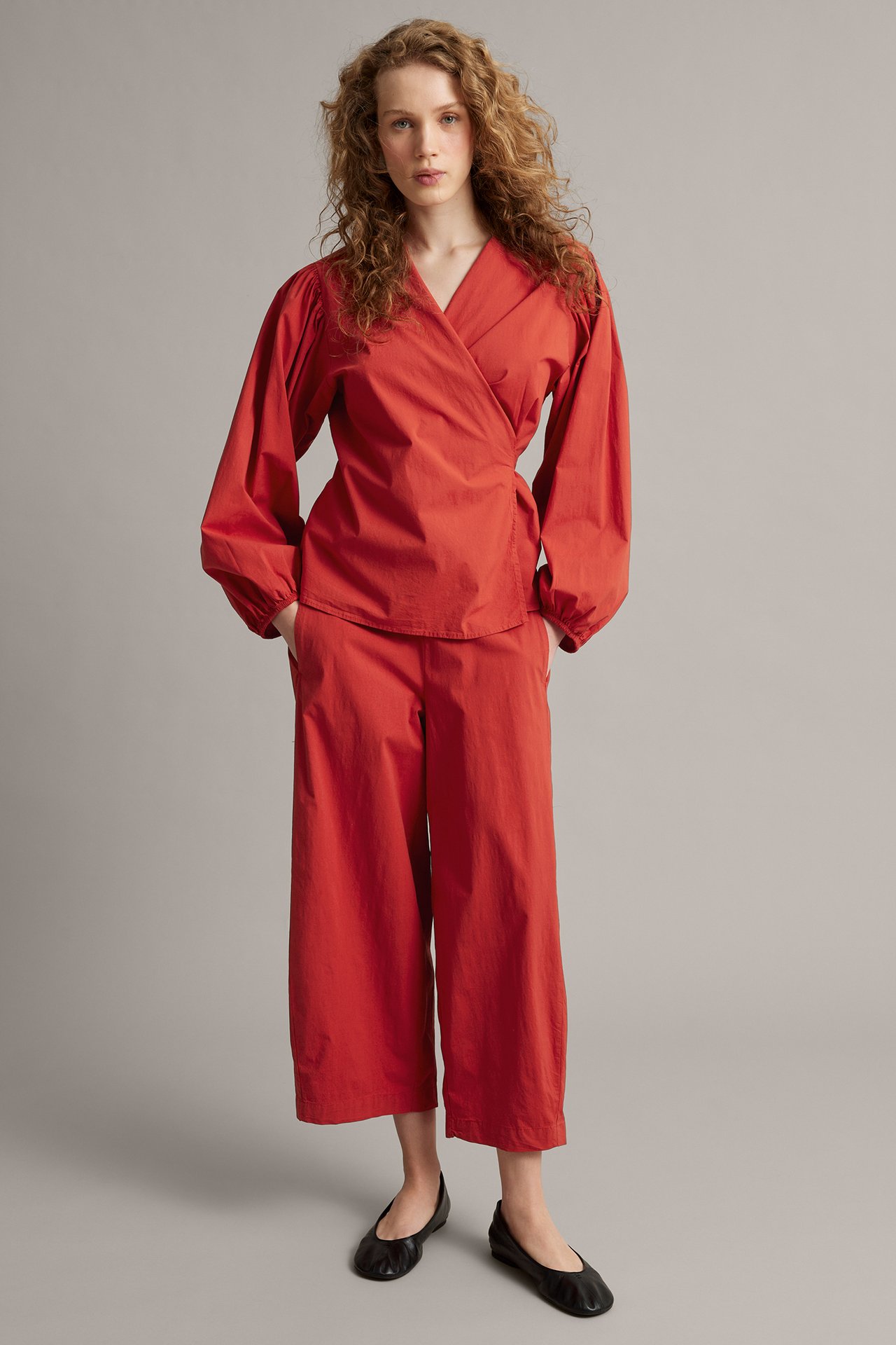 Widok z przodu kobiety w czerwonych, skróconych spodniach barrel-fit, pasującym czerwonym topie kopertowym i czarnych baletkach.