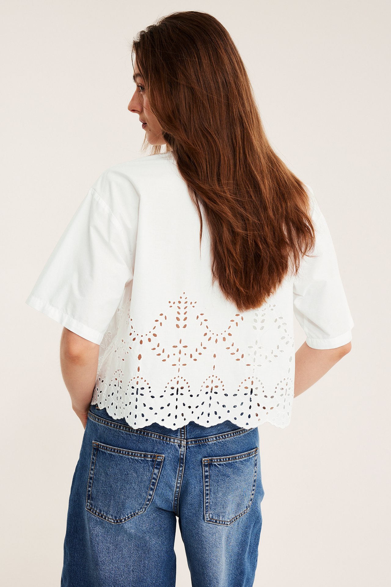 Nainen takaapäin valkoisessa lyhythihaisessa broderie anglaise -paidassa ja sinisissä leveälahkeisissa farkuissa.