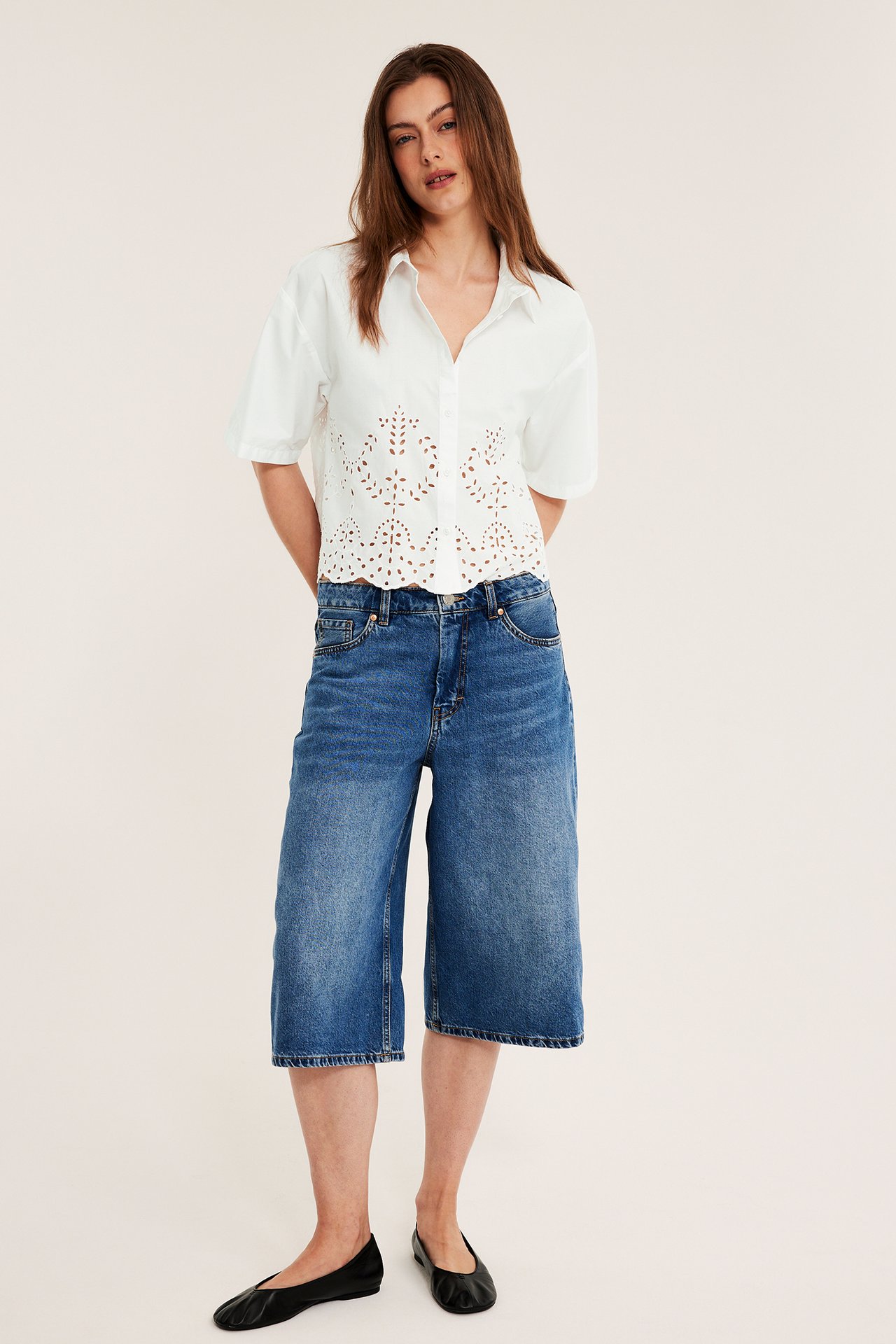 Framifrån, kvinna i vit kortärmad skjorta med broderie anglaise, blå denim culottes, svarta skor.