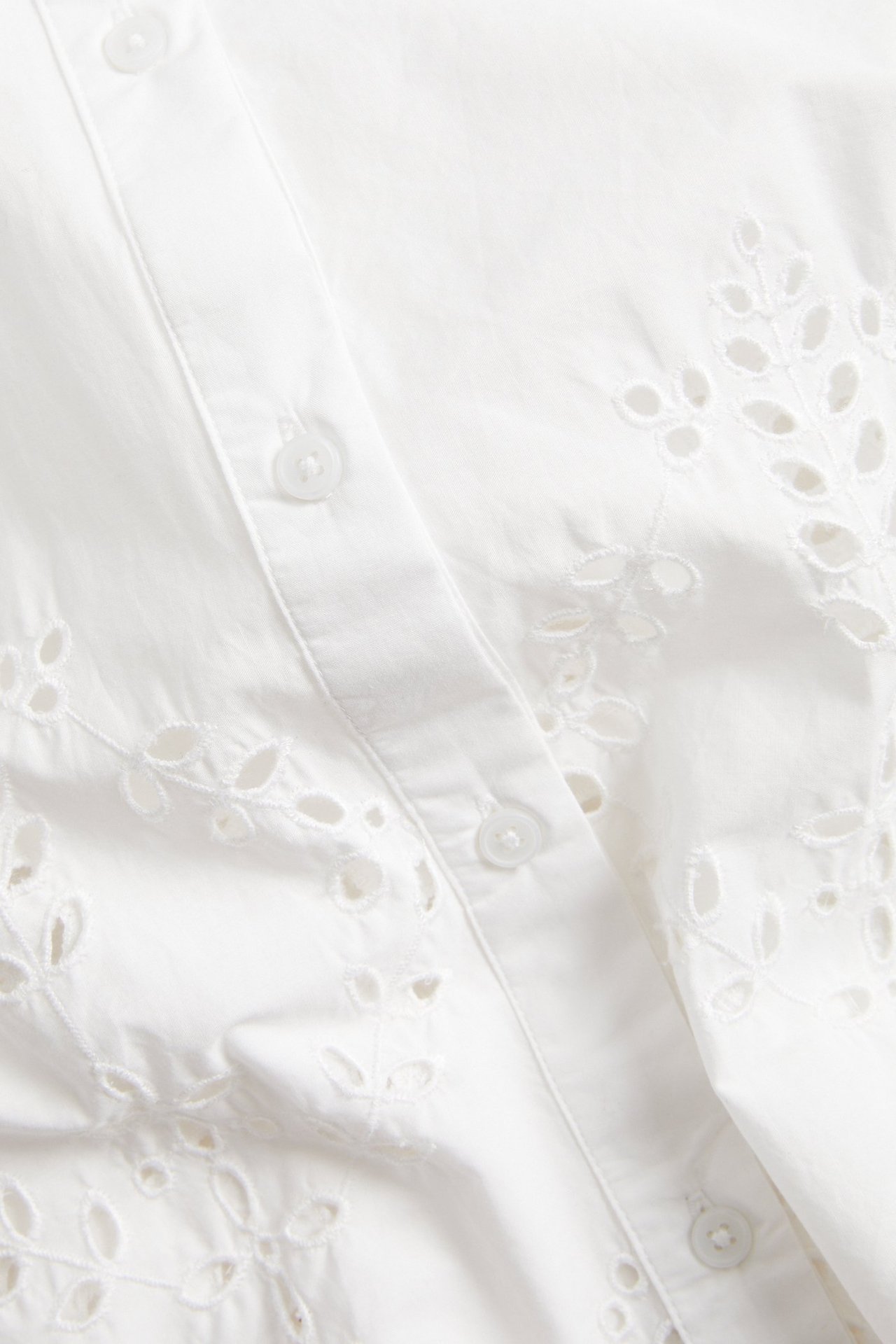 Nærbilde av en hvit kortermet bomullsskjorte med broderie anglaise og knapper foran.