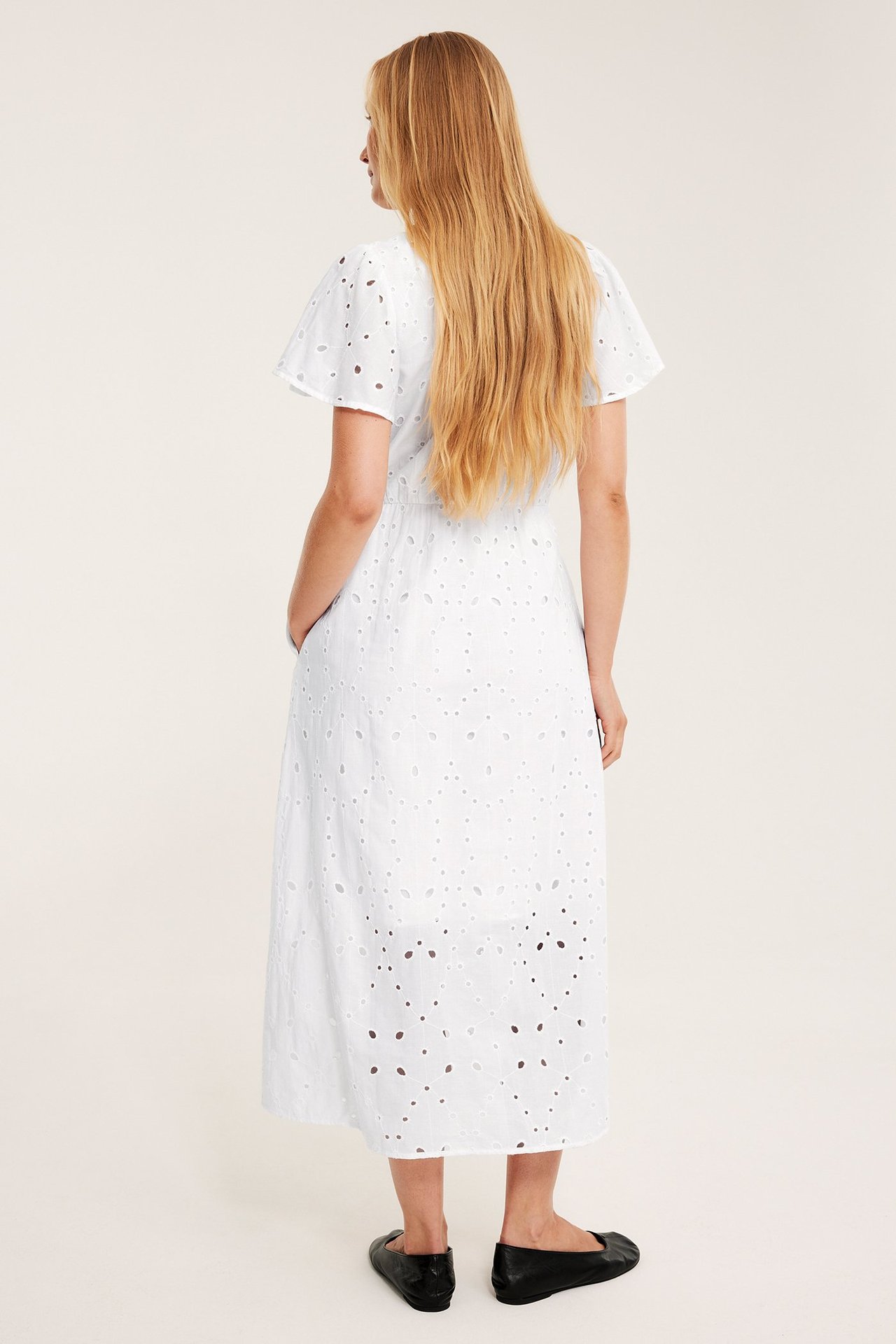 Nainen takaa: valkoinen puuvillainen broderie anglaise -midimekko ja mustat ballerinat.