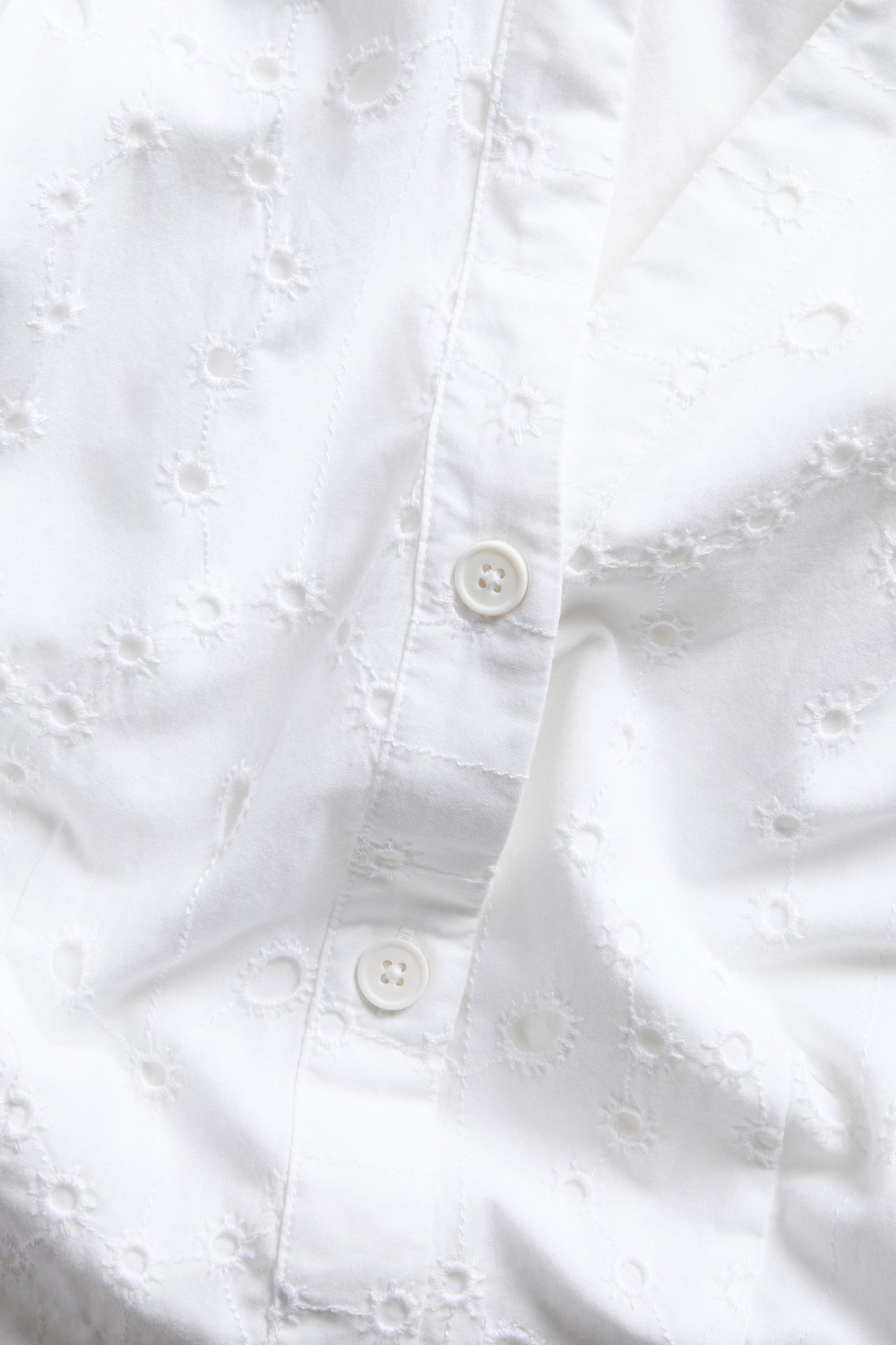 Närbild av en vit bomullsklänning med broderie anglaise-mönster och vit knappslå.