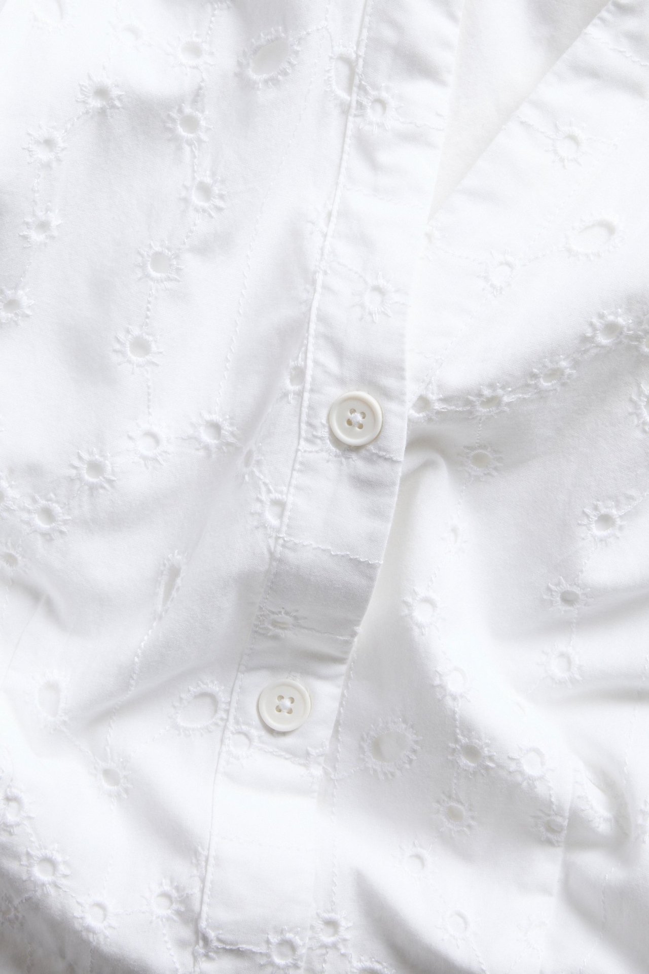 Närbild av en vit bomullsklänning med broderie anglaise-mönster och vit knappslå.