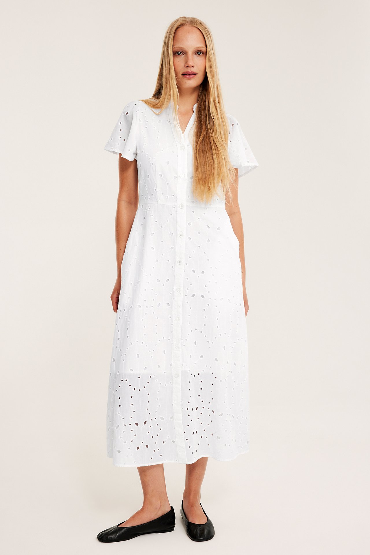 Frontvy av en kvinna i en vit midiklänning med broderie anglaise, korta ärmar och svarta ballerinaskor.