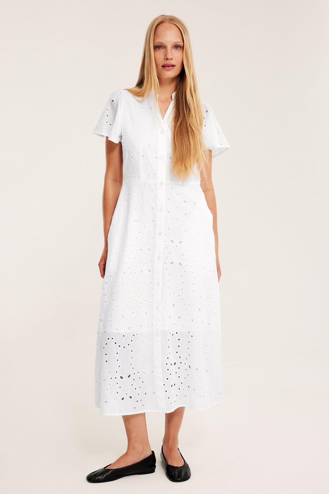 Frontvy av en kvinne i en hvit midikjole med broderie anglaise, korte ermer og svarte ballerinasko.
