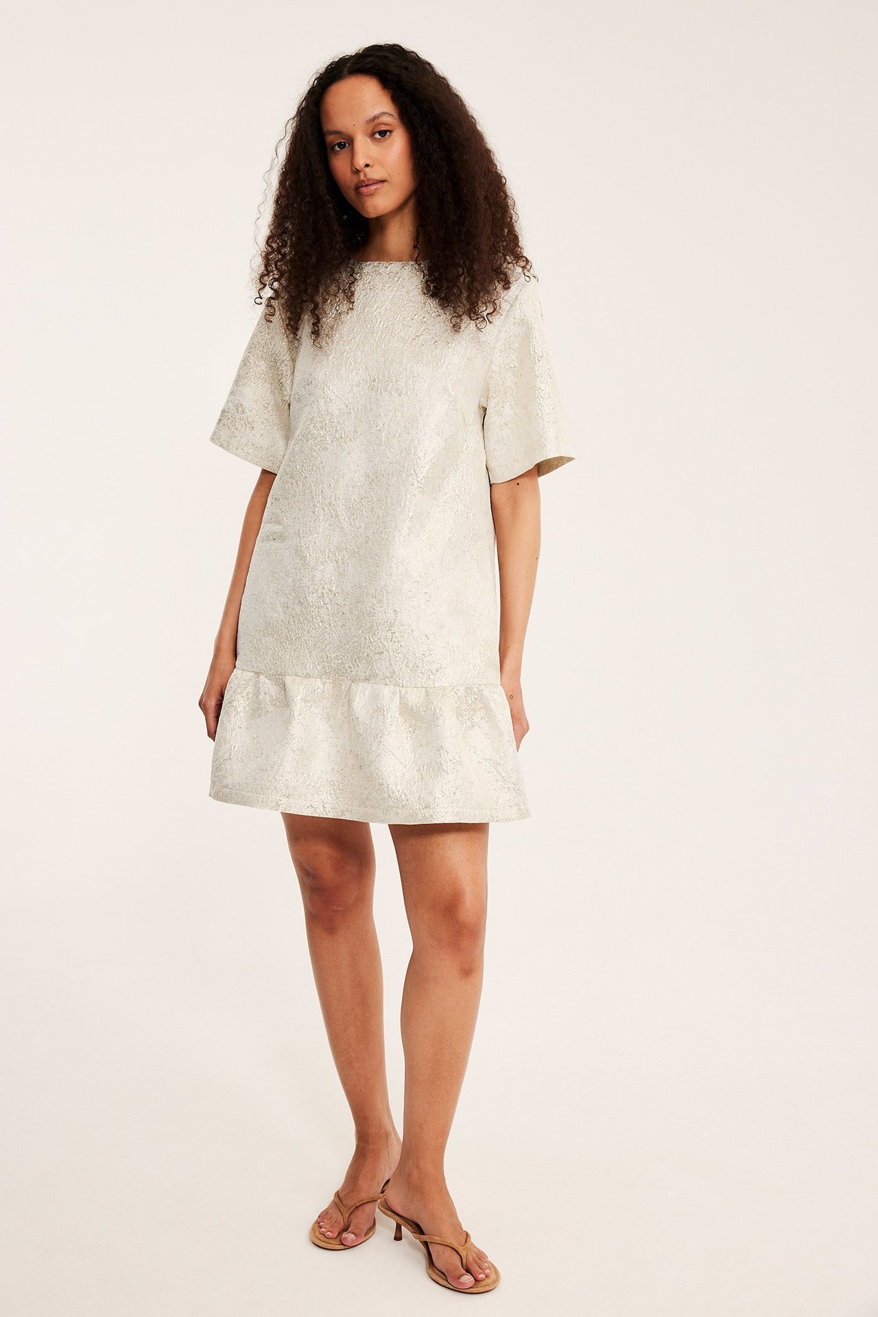 Frontvy av kvinna i offwhite jacquardmönstrad klänning med volangkant och beiga högklackade sandaler.