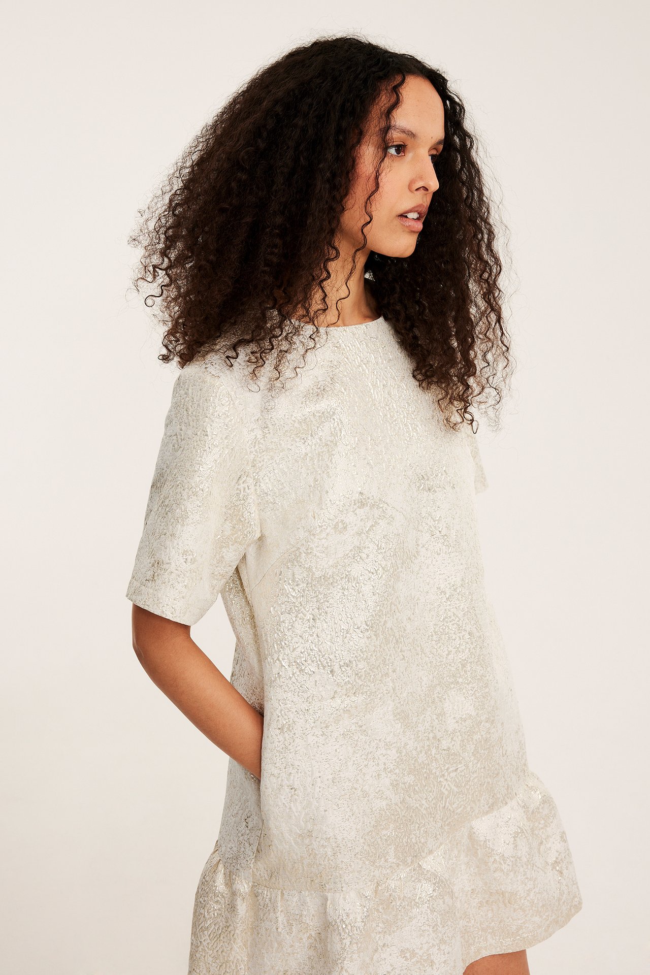 Sidovy av en kvinna i en offwhite jacquardmönstrad klänning med korta ärmar och volangkant.