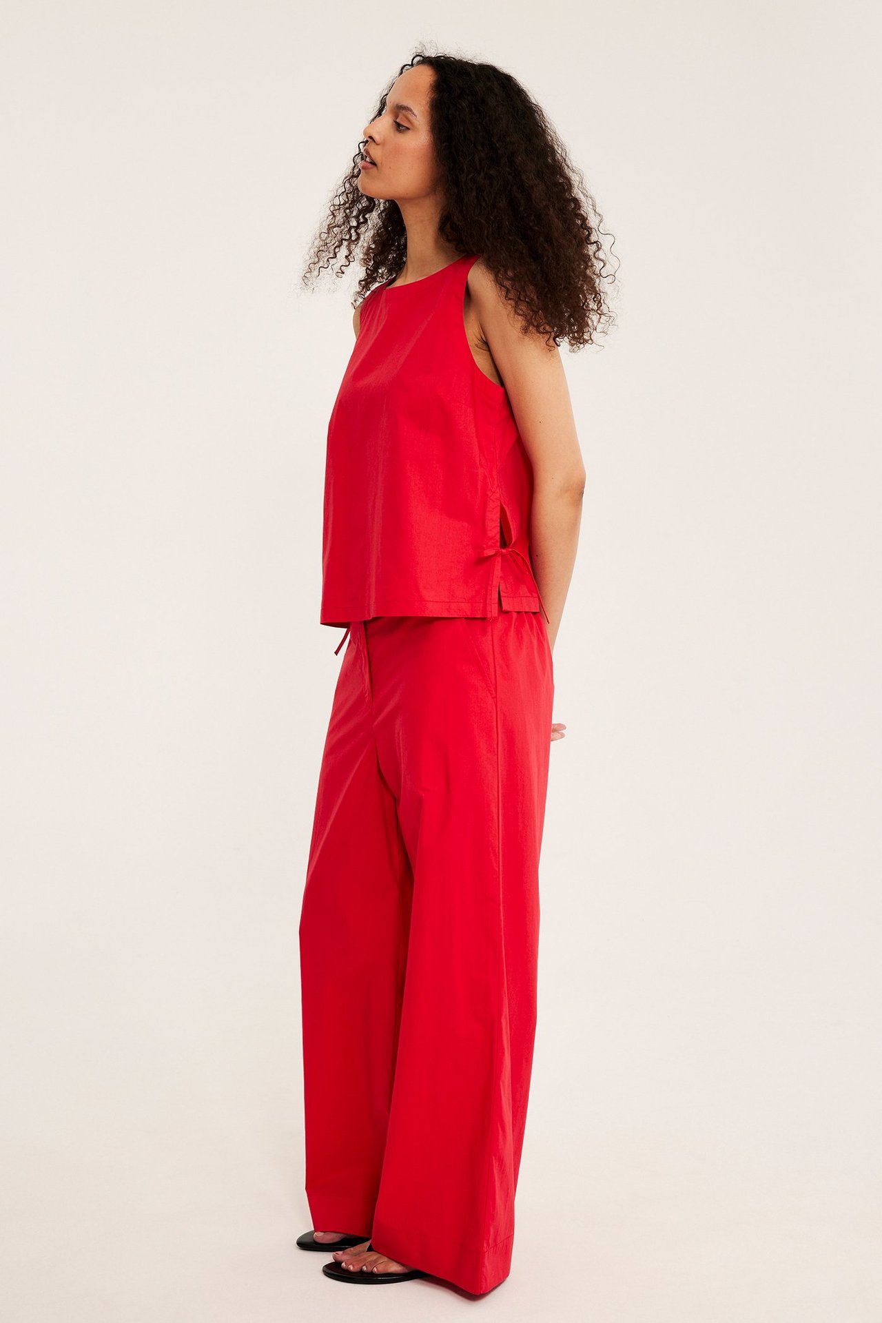 Sidovy av en kvinna i röd ärmlös bomullstopp med knytband i sidan och matchande röda vida byxor.