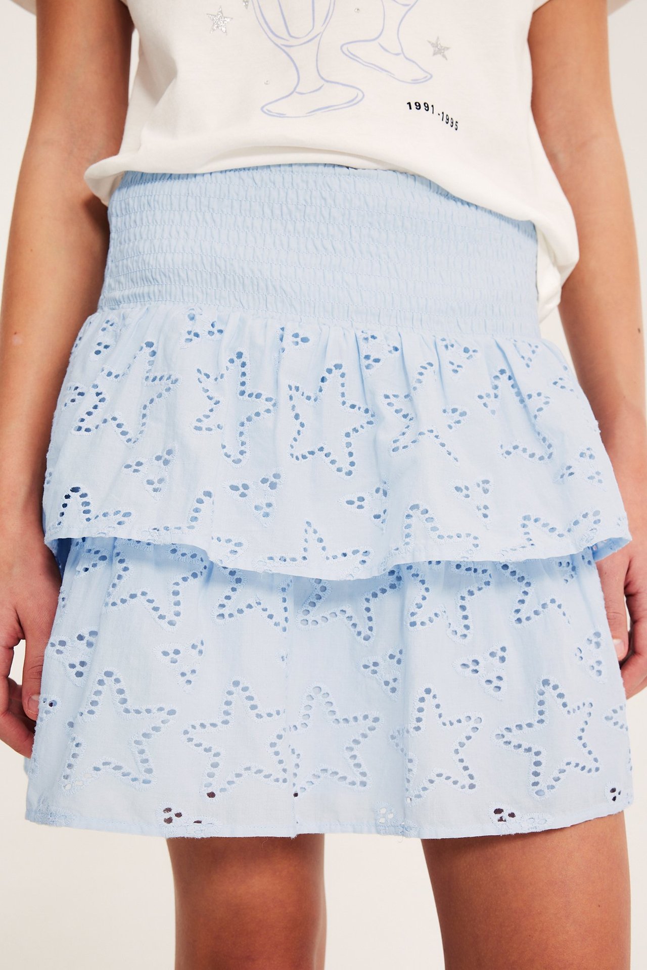 Frontalvisning: Barn bærer lyseblått volanskjørt med stjernebroderi, smocket midje, over hvit t-skjorte.