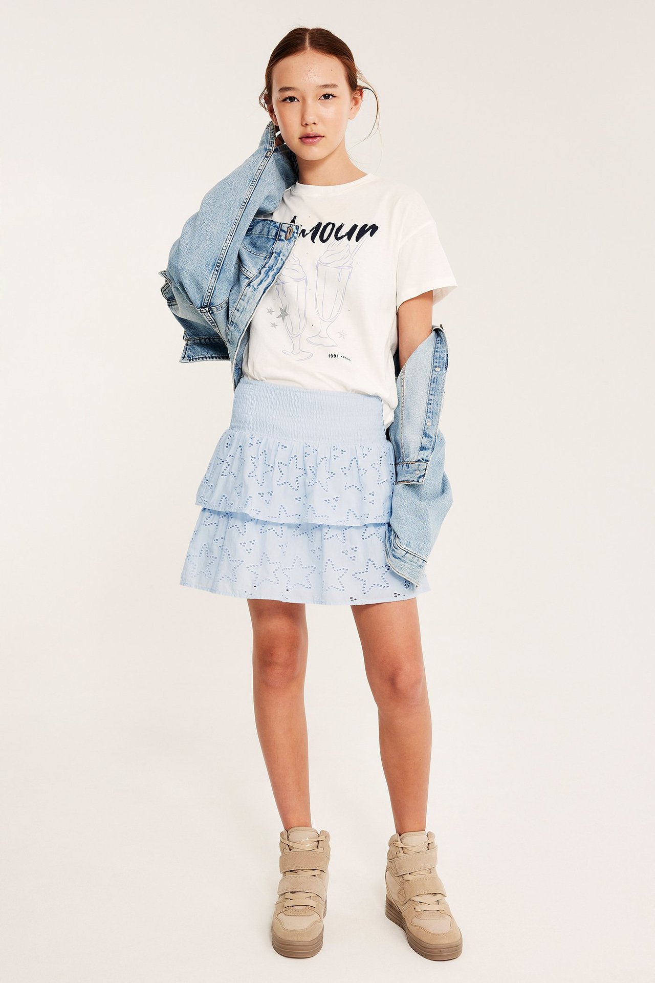 Dziecko nosi jasnoniebieską marszczoną spódnicę z falbankami i haftem w gwiazdki, biały t-shirt, kurtkę jeansową, beżowe trampki.