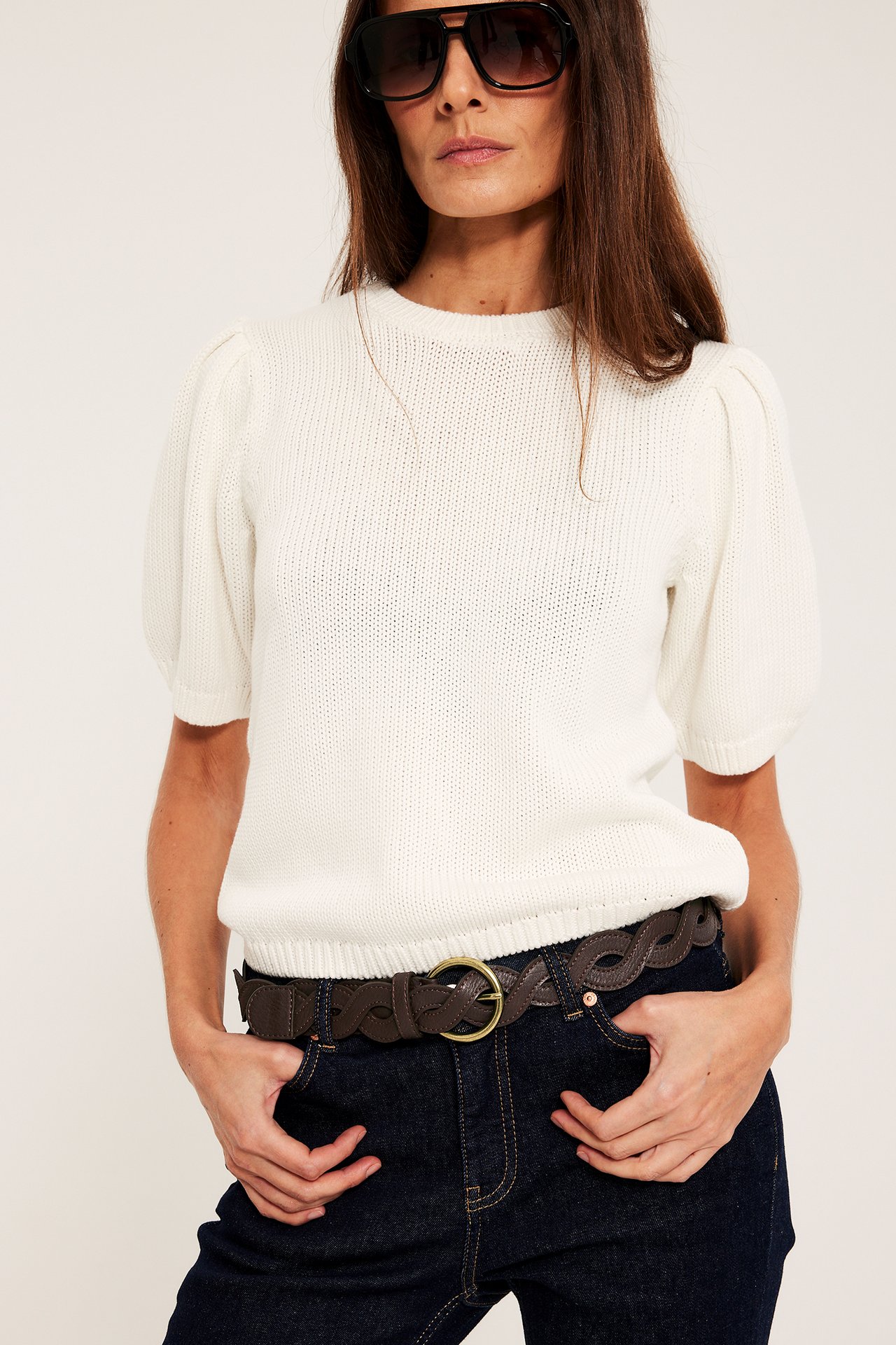 Forfra: Kvinne med brunt flettet belte, gullspenne, hvit strikket topp, mørke jeans, svarte solbriller.