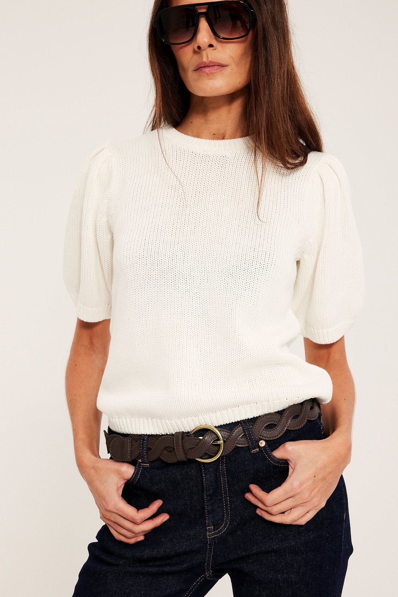 Edestä: Nainen yllään ruskea punottu vyö, kultasolki, valkoinen neuletoppi, tummat farkut, mustat aurinkolasit.