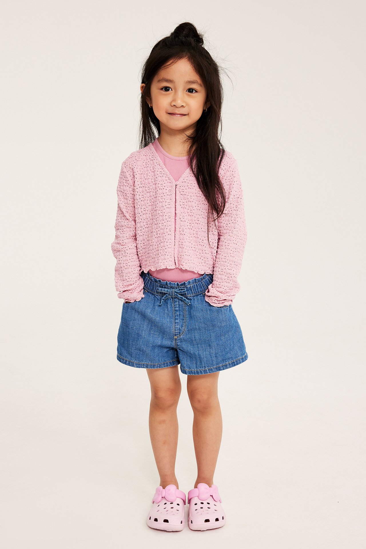 Framifrån: barn i ljusrosa spetskofta över rosa topp, blåa jeansshorts och rosa tofflor.