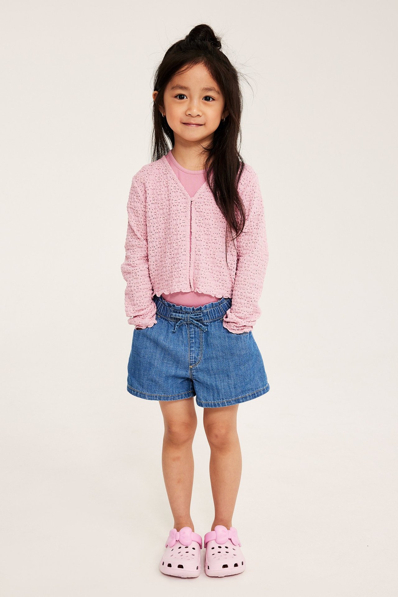 Edestä: lapsi yllään vaaleanpunainen pitsineuletakki, vaaleanpunainen toppi, siniset denim shortsit ja vaaleanpunaiset sandaalit.