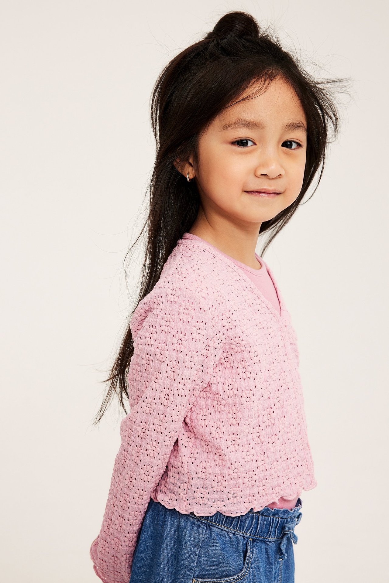 Etunäkymä: Lapsi yllään ohut vaaleanpunainen pitsineuletakki pinkin topin ja sinisten farkkushortsien päällä.