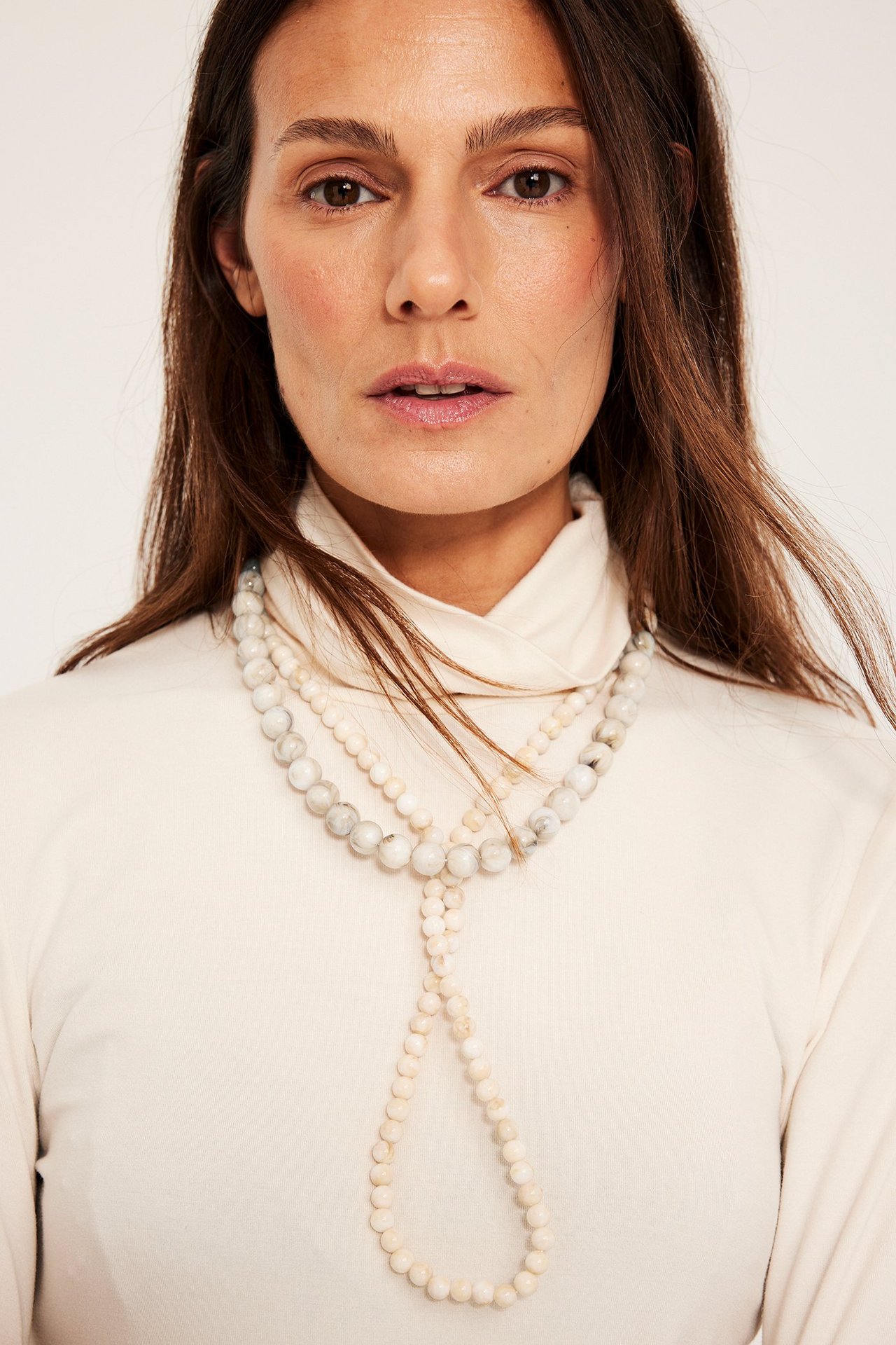 Närbild av kvinna som bär lager på lager av ljusbeige och gråaktiga pärlhalsband över en beige polotröja.