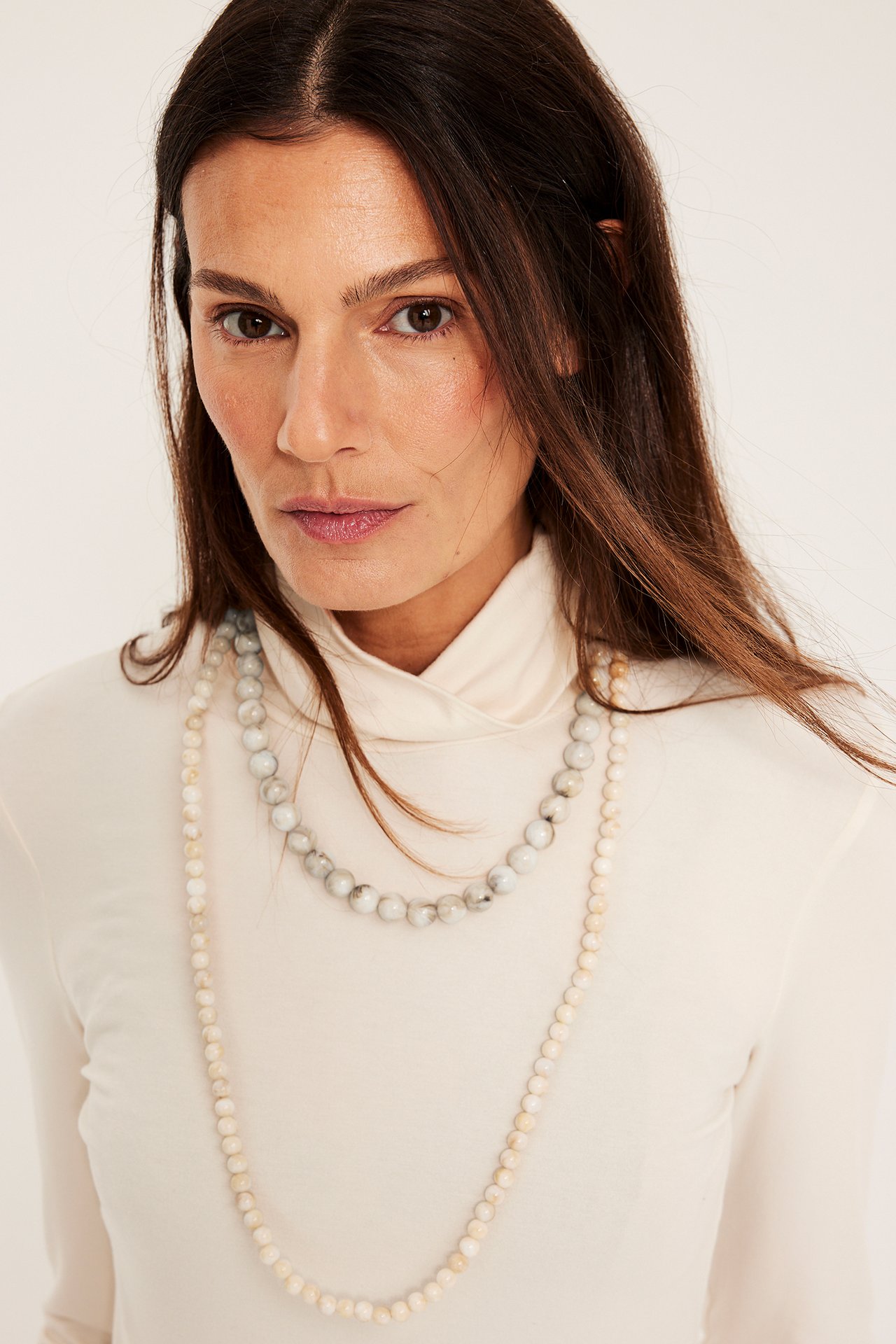 Långt ljusbeige pärlhalsband och ett gråmelerat halsband, burna av kvinna över en beige polotröja, närbild.