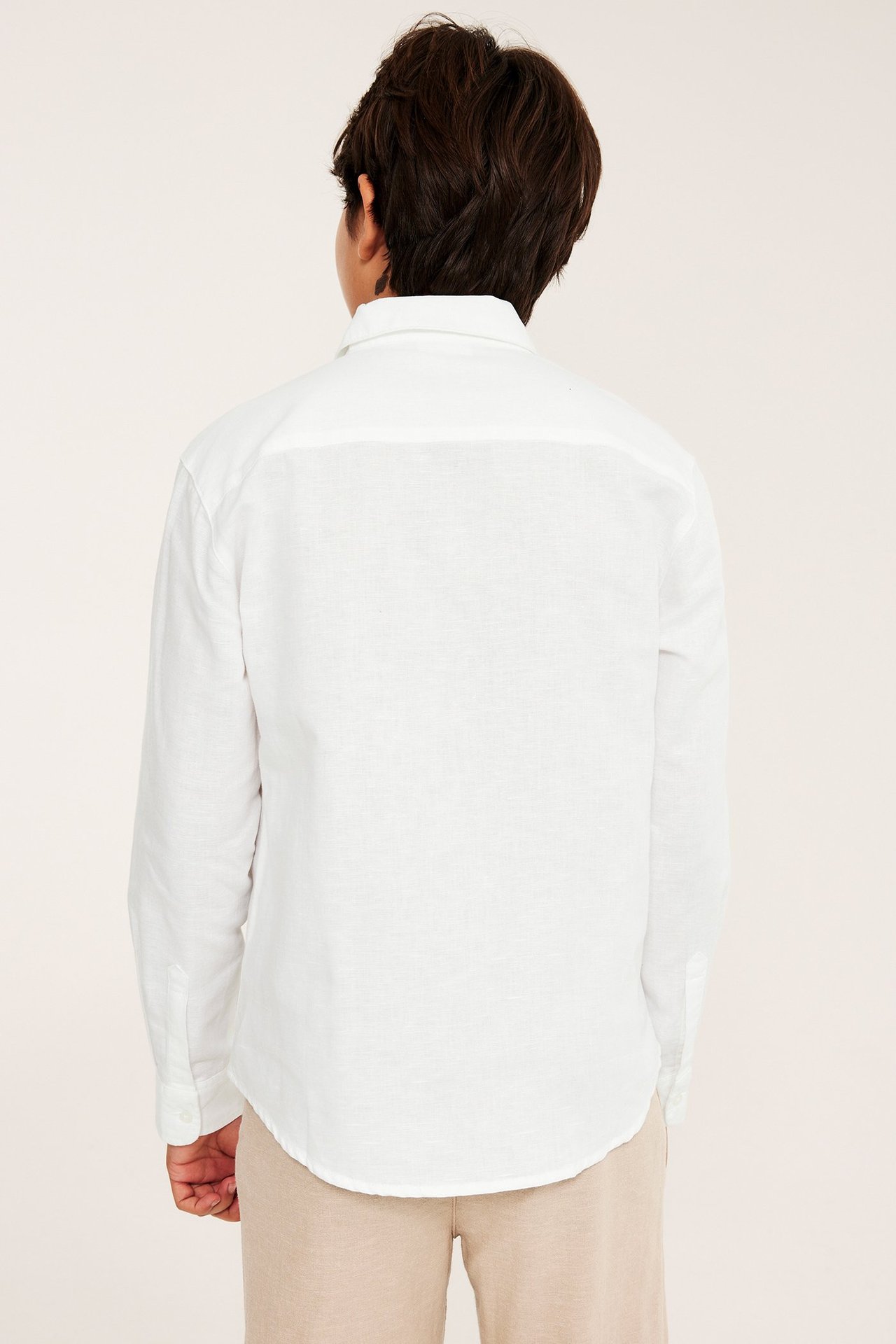 Bakfra av et barn som har på seg en hvit langermet skjorte i linblanding og beige bukser.