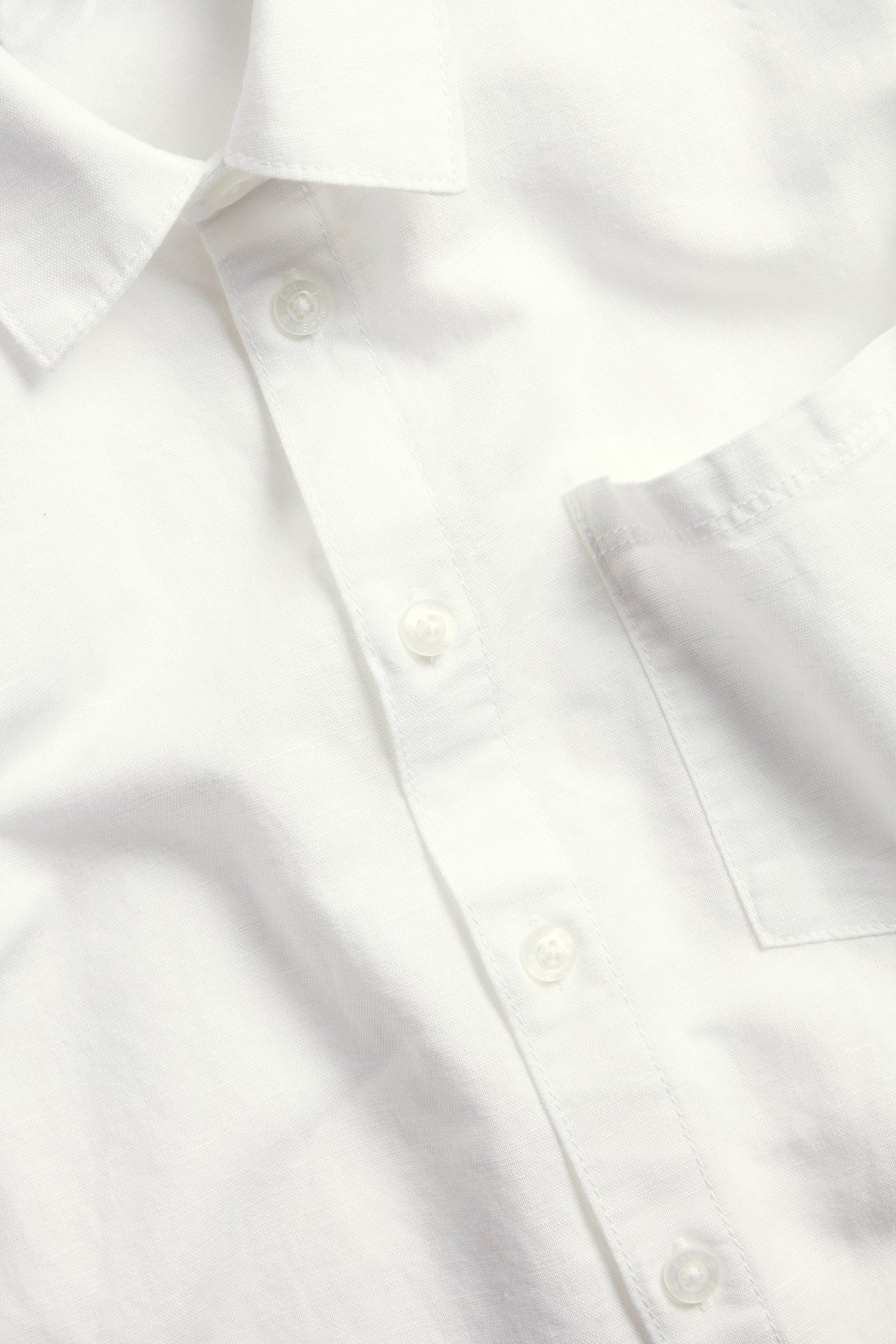 Närbild på en vit linneblandningsskjorta med krage, knappslå och bröstficka.