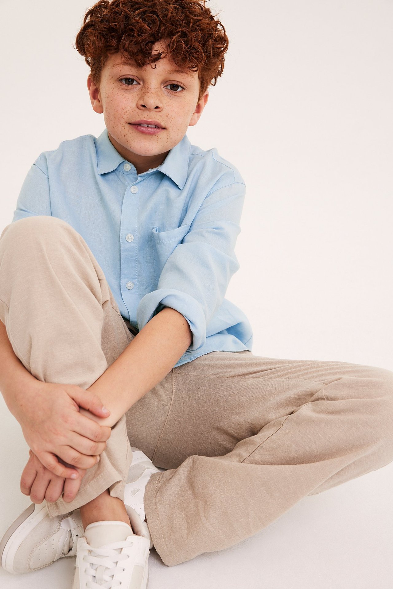 Dziecko w jasnoniebieskiej koszuli z lnu, podwinięte rękawy, beżowe spodnie i białe trampki.