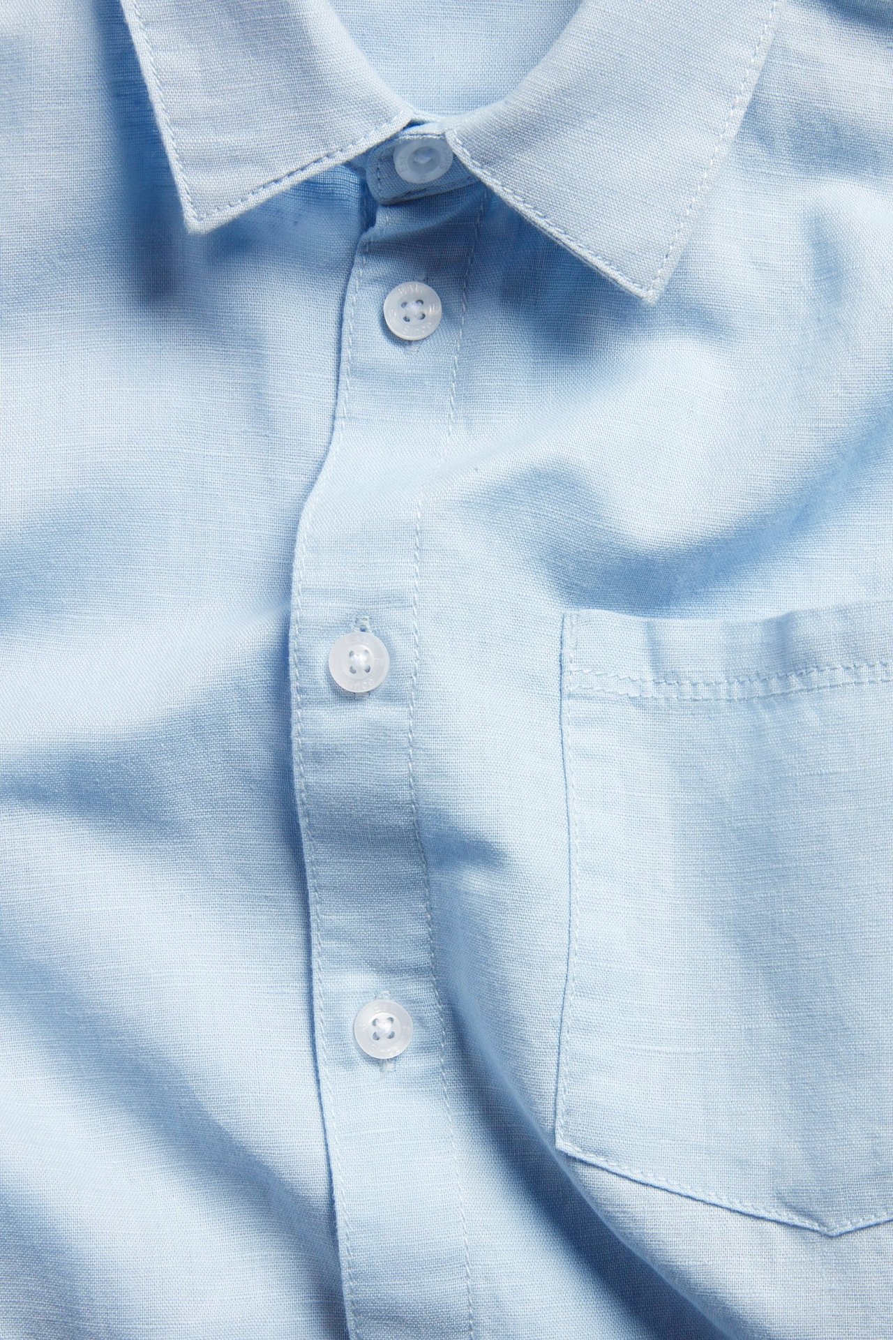 Närbild av en ljusblå skjorta i linnemix med vita knappar och en bröstficka.