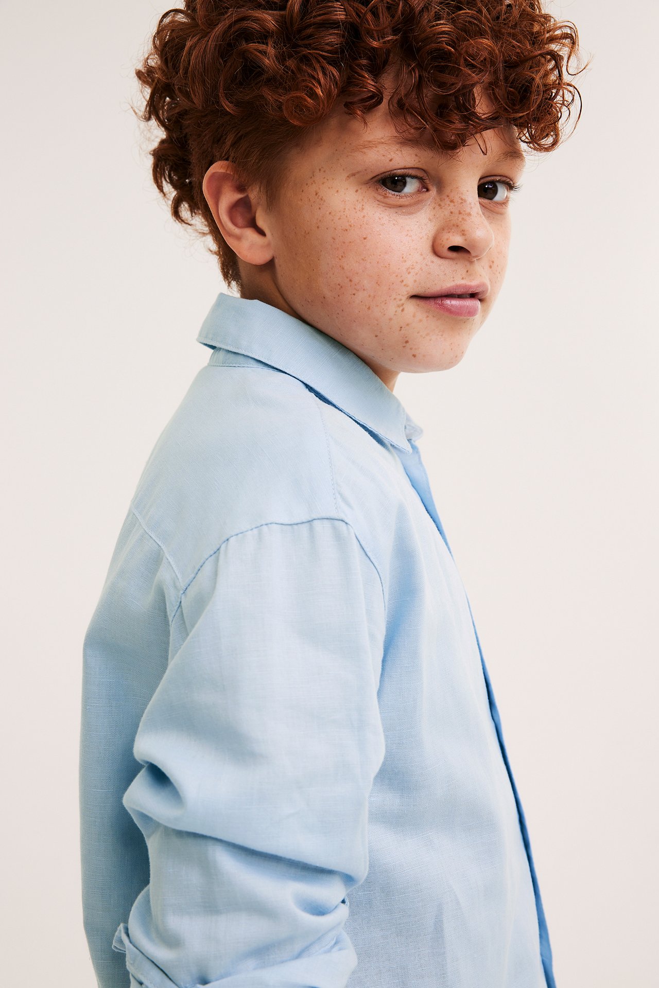 Närbild, sidovy av pojke i ljusblå enfärgad skjorta i linnemix med upprullade ärmar.