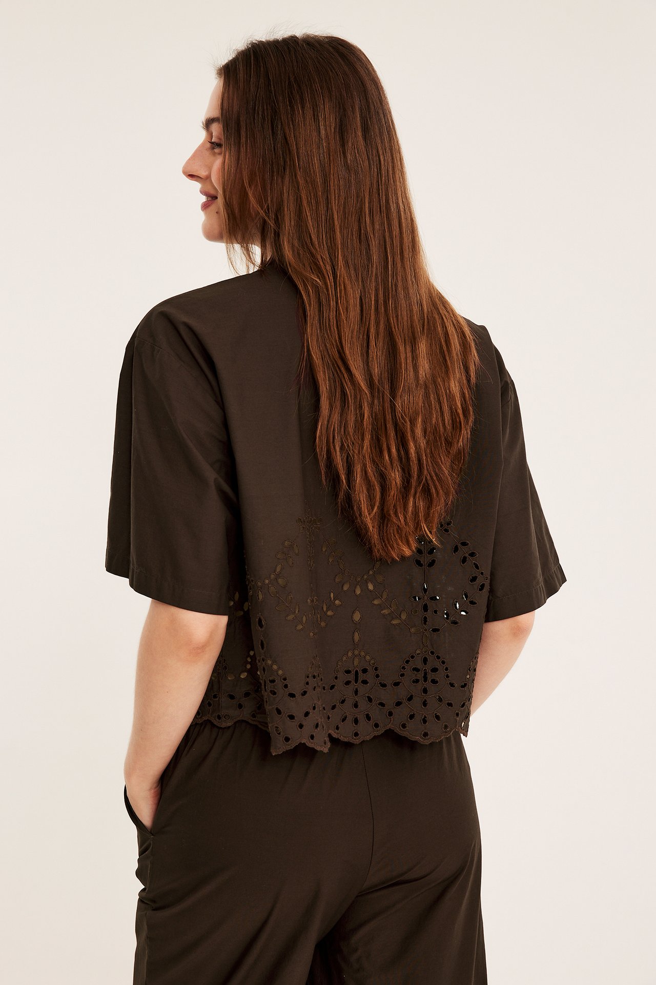 Naisen sivu/selkäkuva tummanruskeassa lyhythihaisessa broderie anglaise -paidassa ja yhteensopivissa housuissa.