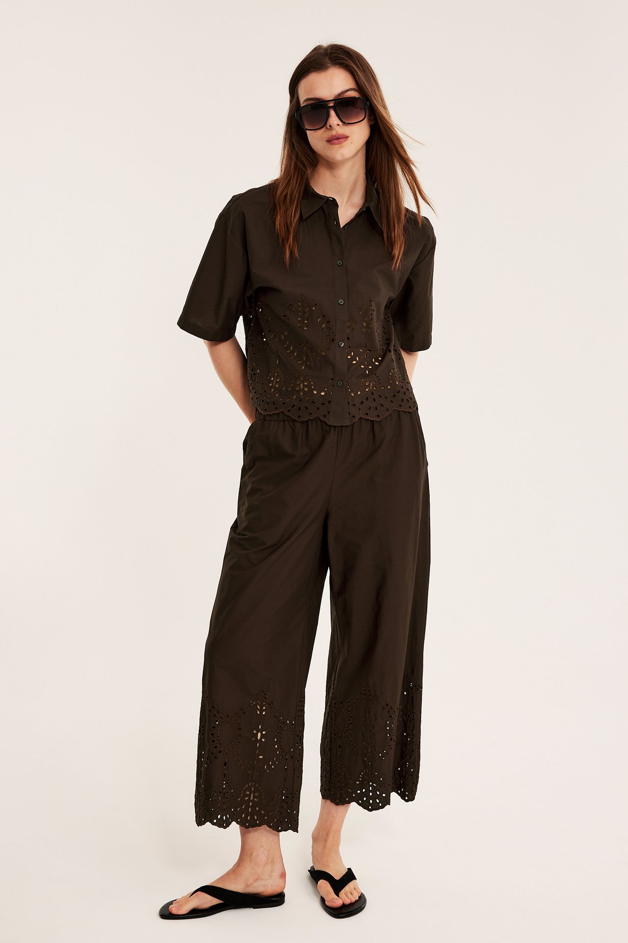 Kvinna i mörkbrun brodyrskjorta, matchande byxor, svarta flip-flops och solglasögon. Framsida.