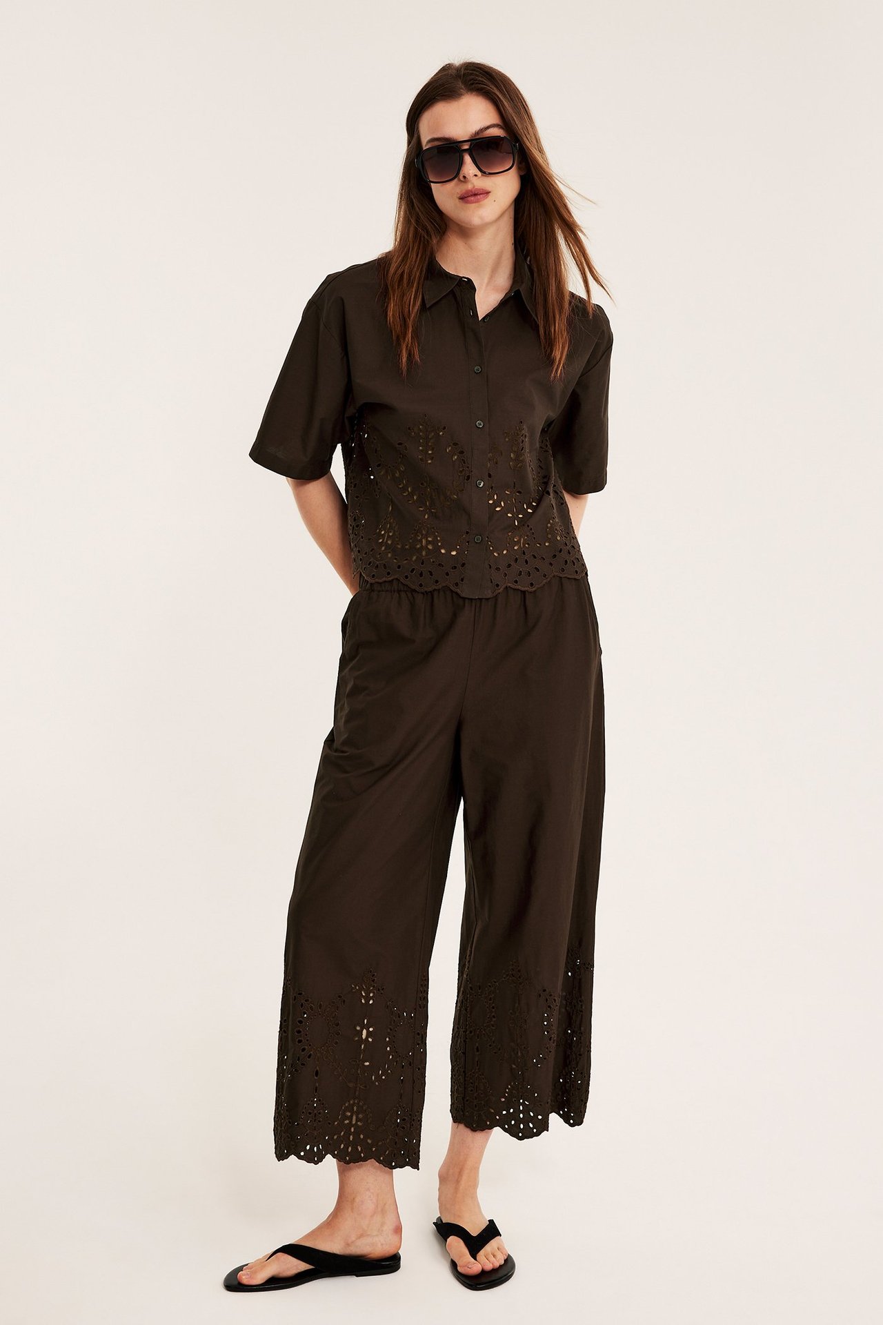 Nainen tummanruskeassa brodyyripaidassa, yhteensopivissa housuissa, mustissa varvassandaaleissa, aurinkolaseissa. Edestä.