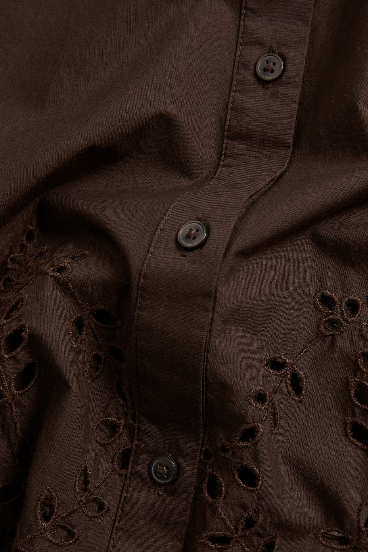 Nærbilde av en brun kortermet bomullsskjorte med knappestolpe og broderie anglaise.