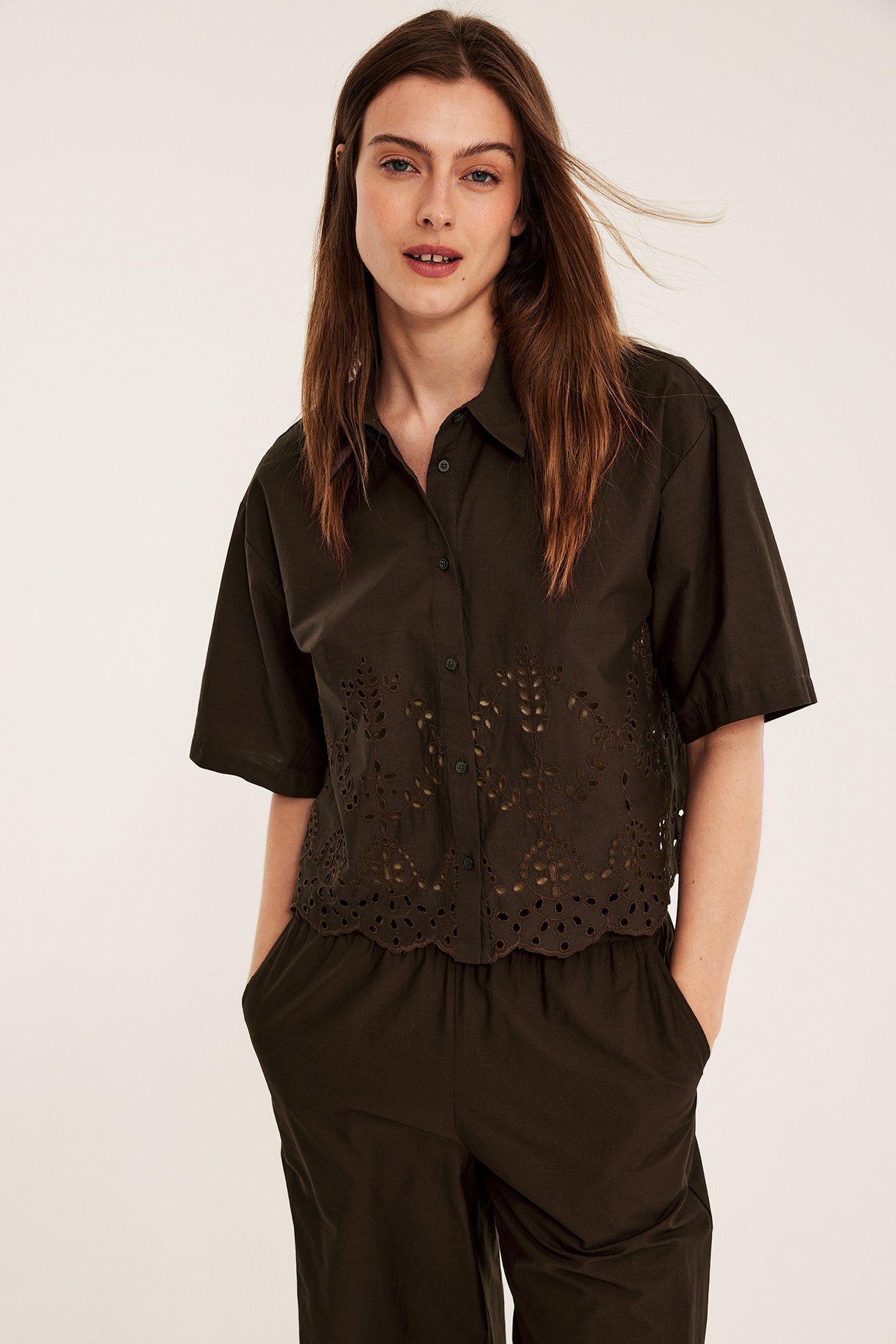 Frontvy av en kvinna i brun kortärmad bomullsskjorta med broderie anglaise och matchande bruna byxor.
