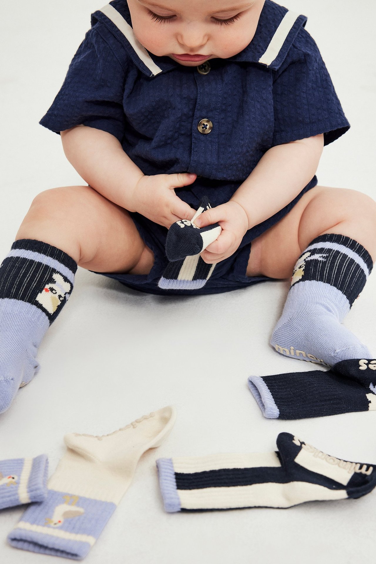 Zbliżenie na dziecko w granatowym kombinezonie i wzorzystych skarpetkach, trzymające pasiastą skarpetę antypoślizgową.