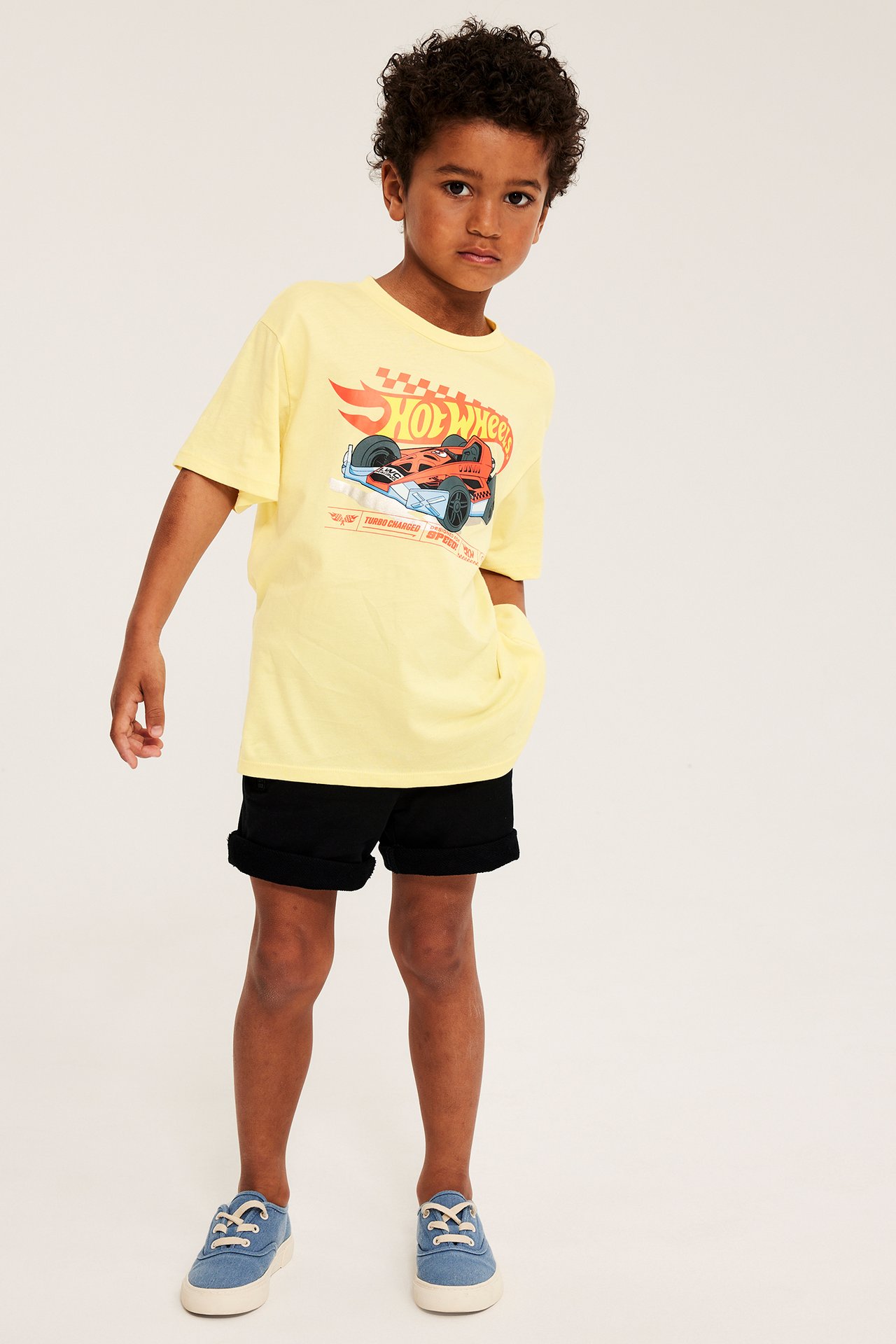 Frontbilde av et barn i en gul bomulls-Hot Wheels t-skjorte, svarte shorts og blå sneakers.