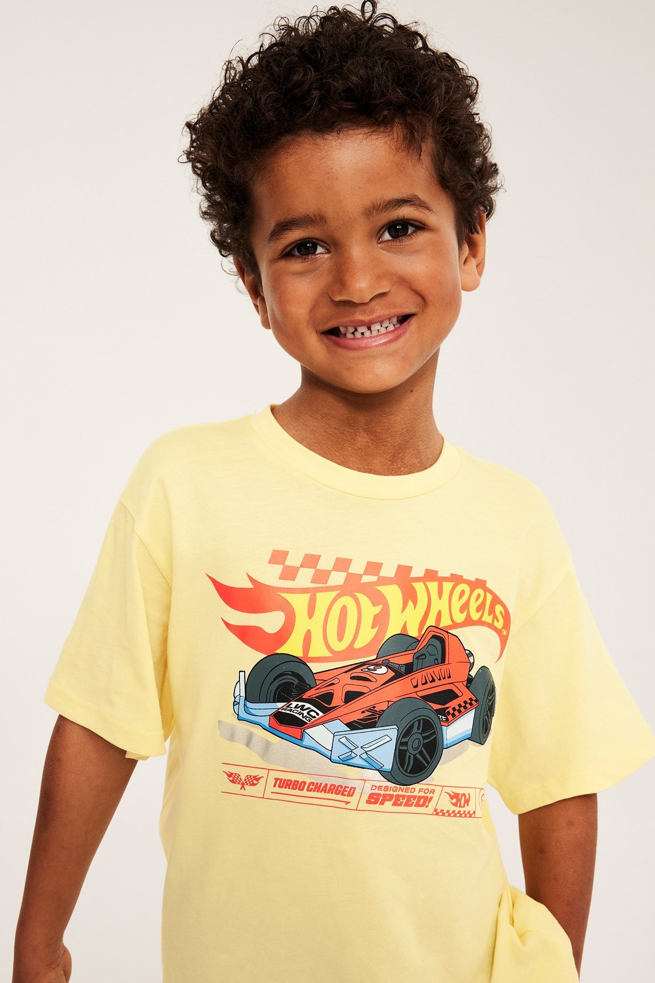 Widok z przodu uśmiechniętego dziecka w żółtym bawełnianym t-shircie Hot Wheels z luźnym krojem.
