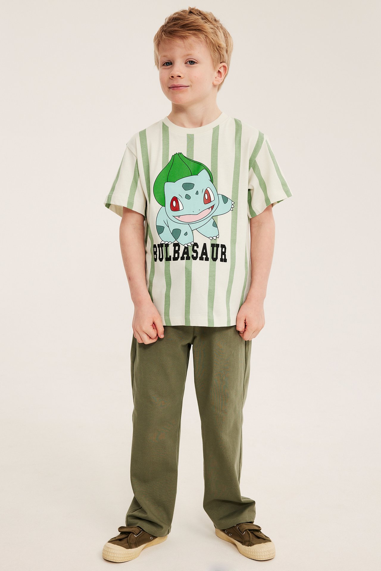 Etunäkymä: poika yllään ylisuuri vaaleanbeige raidallinen Bulbasaur-T-paita, oliivinvihreät housut ja tennarit.