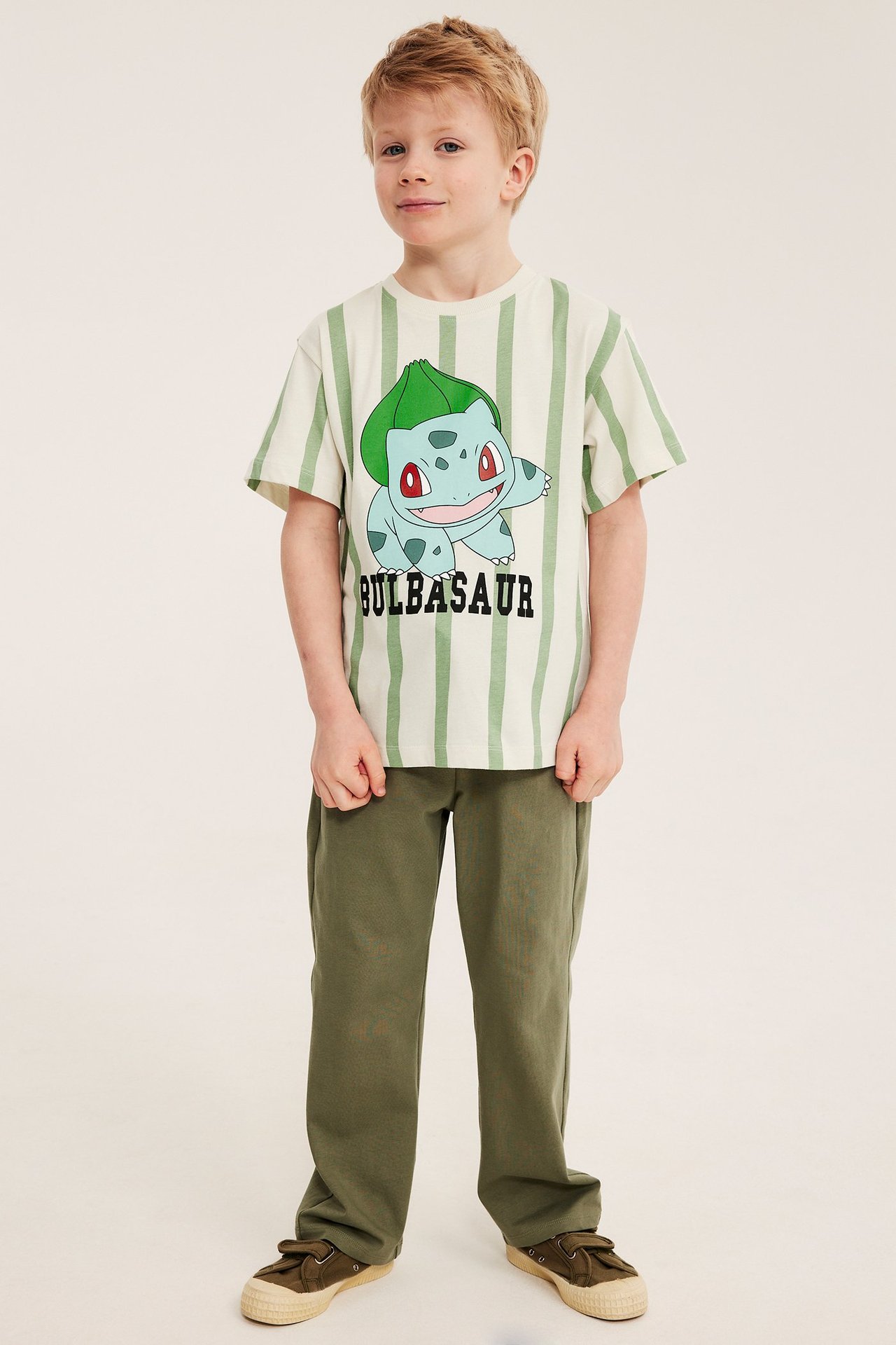 Etunäkymä: poika yllään ylisuuri vaaleanbeige raidallinen Bulbasaur-T-paita, oliivinvihreät housut ja tennarit.