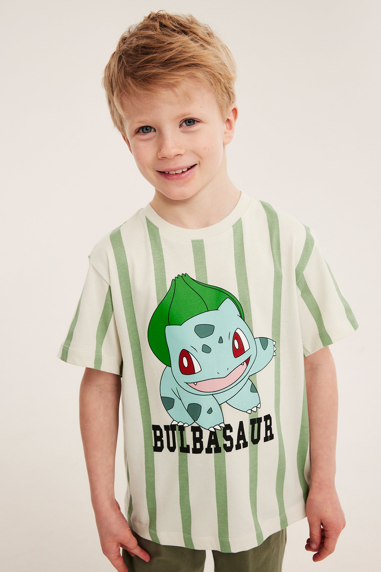 Frontvy av ett barn i en överdimensionerad randig beige t-shirt med Bulbasaur-tryck och gröna byxor.