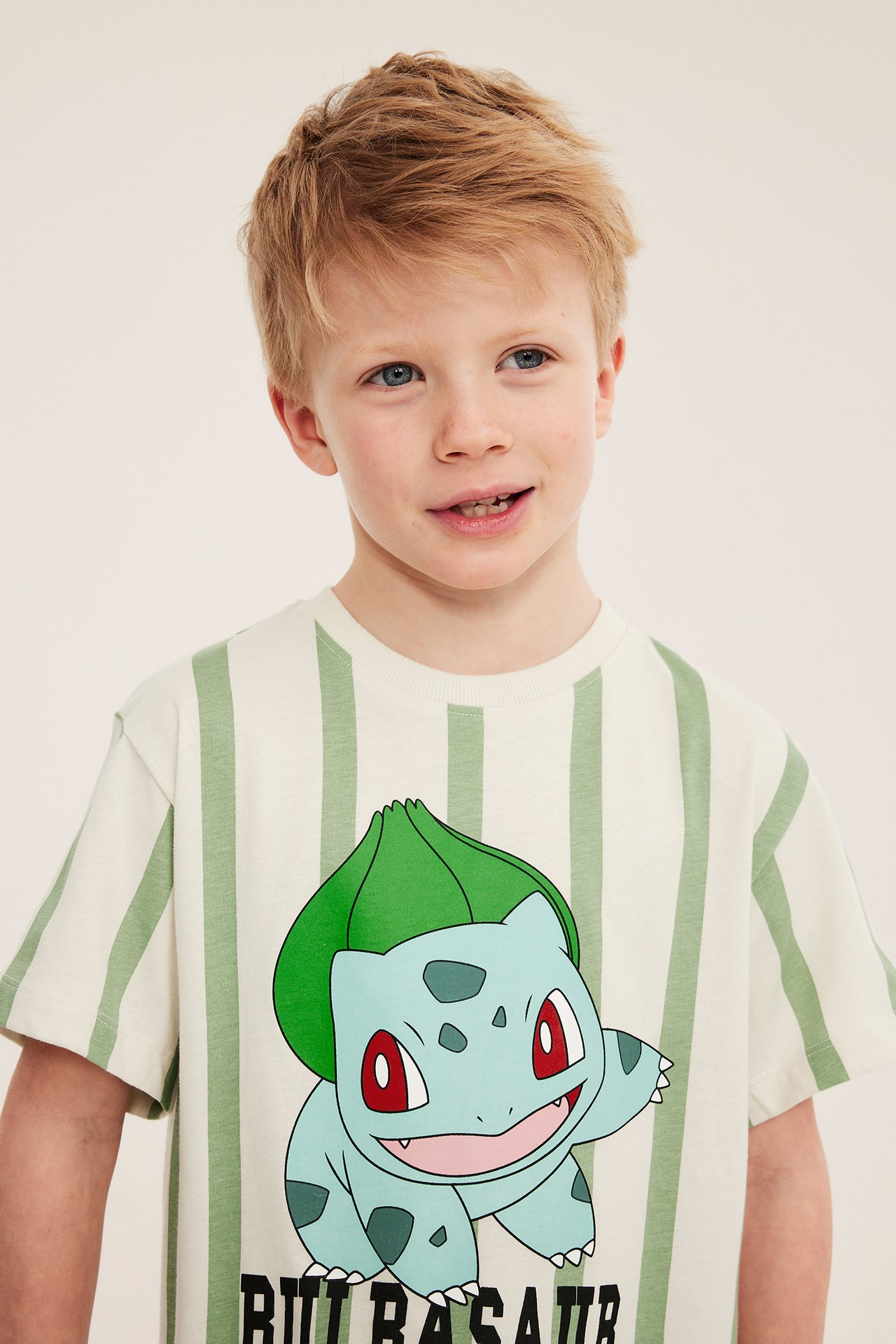 Edestä, lapsi vaaleanbeige-vihreässä raidallisessa Bulbasaur-printti T-paidassa puuvillaa.