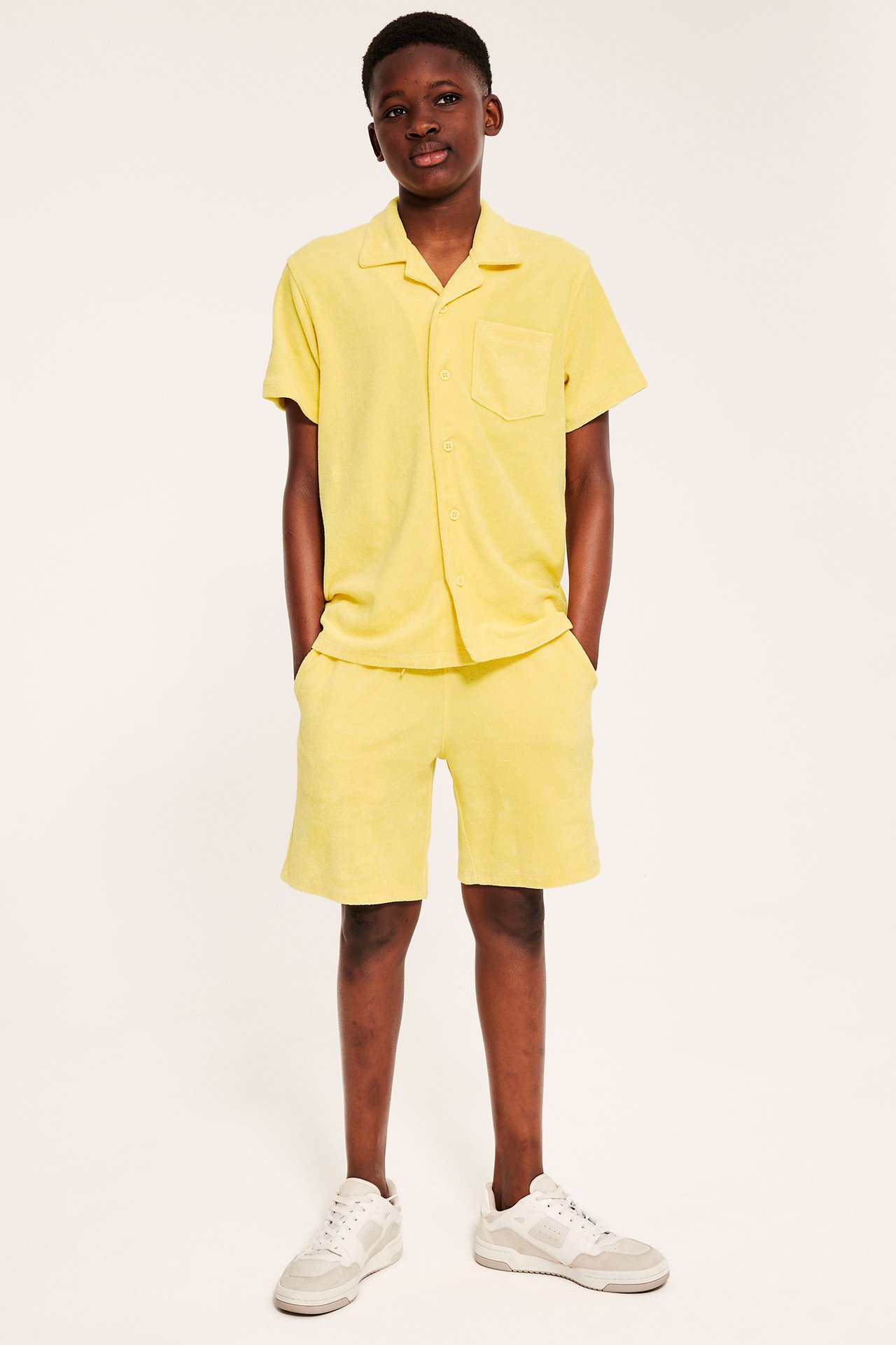 Dziecko w żółtej krótkiej koszuli frotte z kołnierzem resort, pasujących szortach i sneakersach.