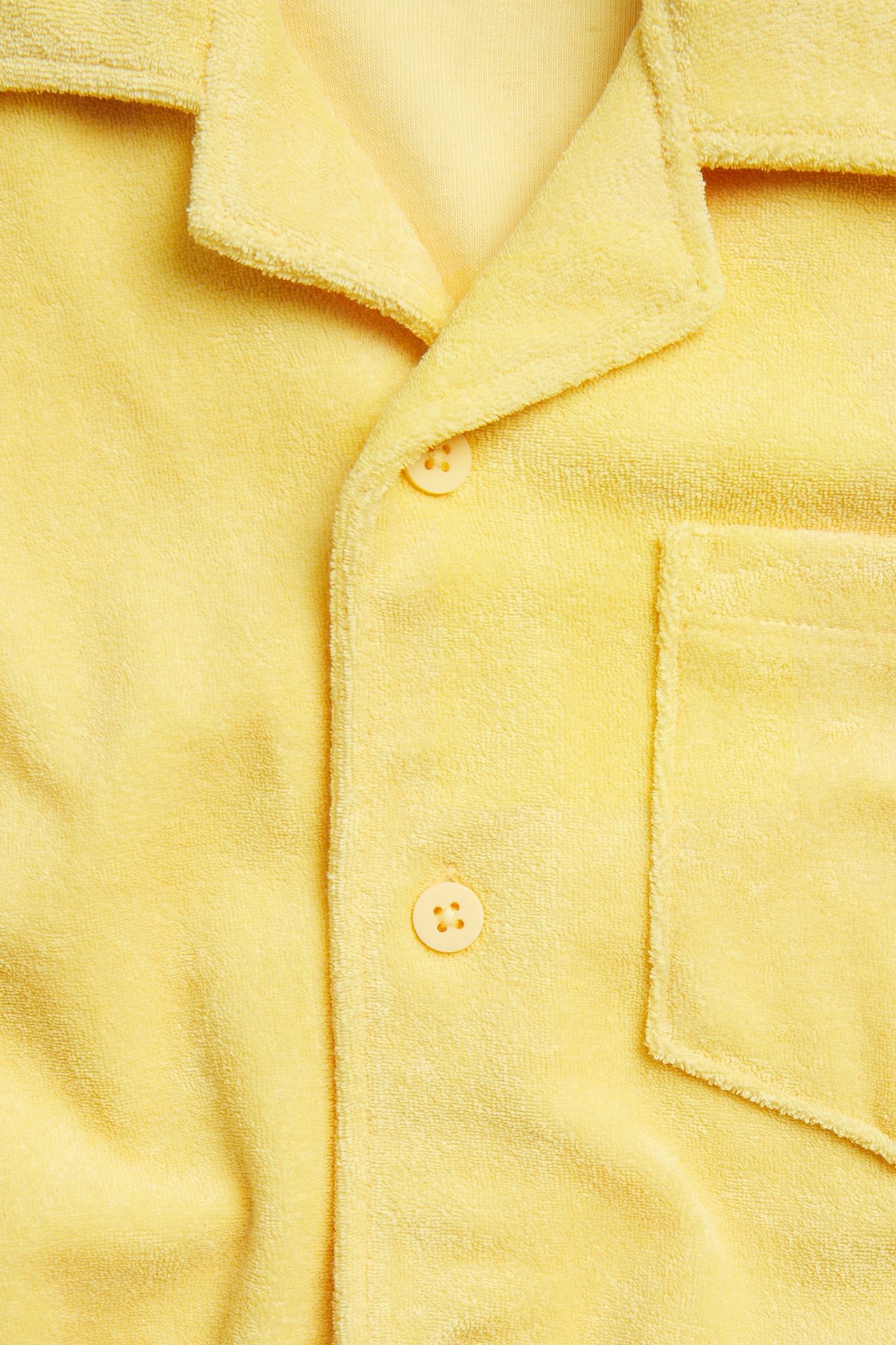 Lähikuva keltaisesta lyhythihaisesta froteepaidasta, jossa on resortkaulus, rintatasku ja etunapit.