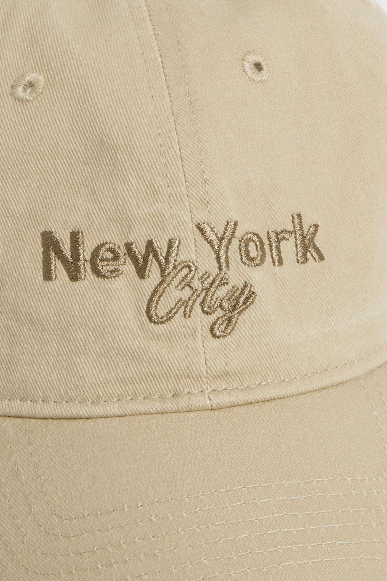 Zbliżenie beżowej czapki z twillu o spranej fakturze, z brązowym haftem 'New York City'.
