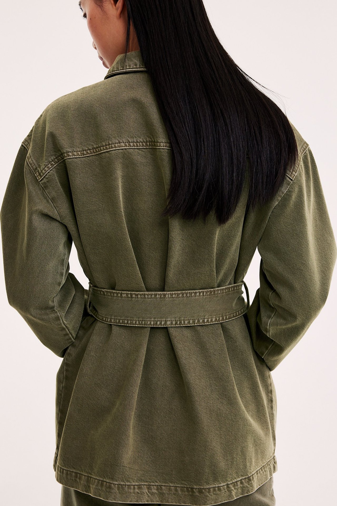 Naisen selkäkuva, yllään väljä vihreä twill-utilitytakki vyöllä vyötäröllä.