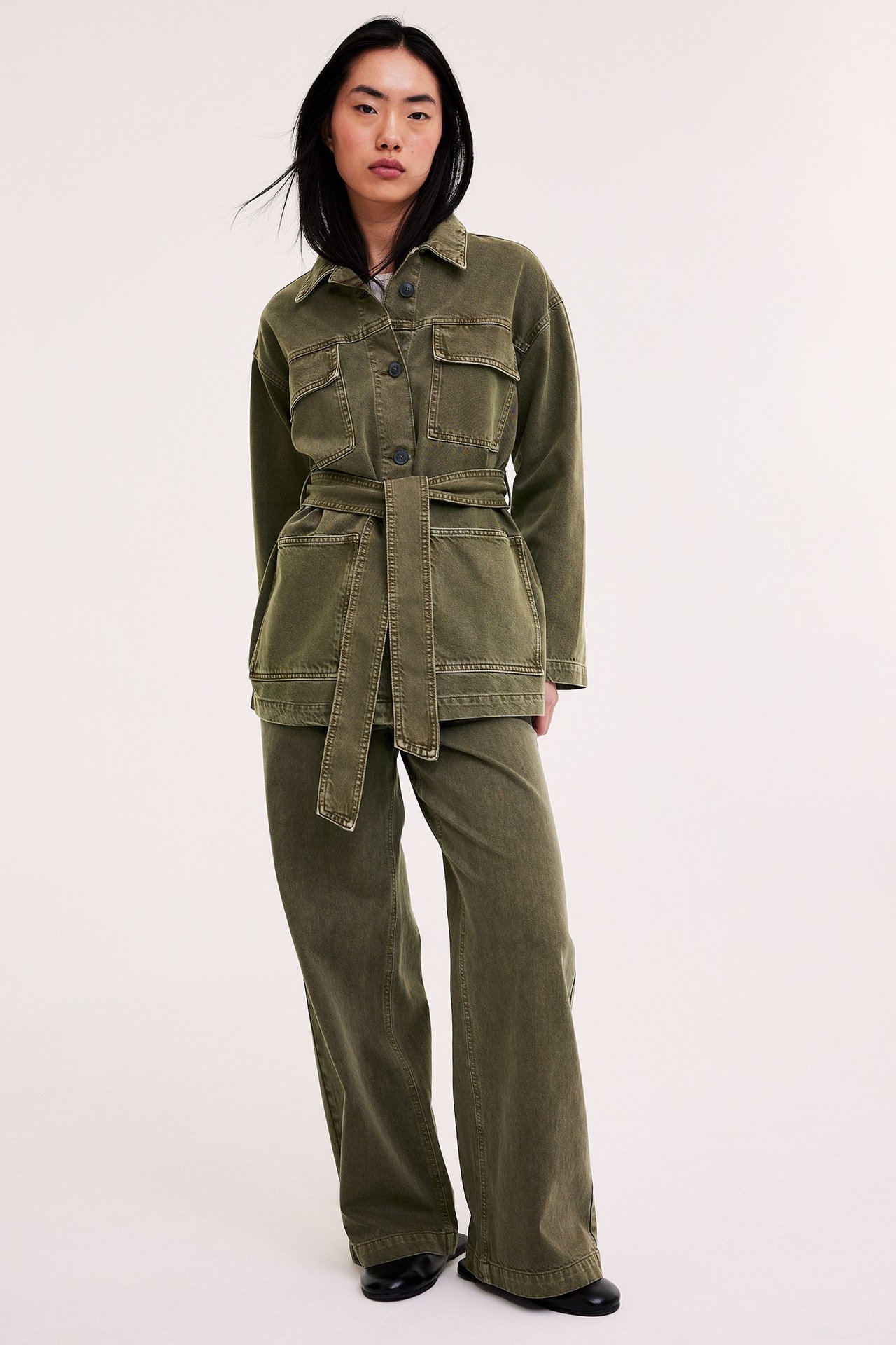 Nainen yllään vyöllinen vihreä puuvillatakki, yhteensopivat leveälahkeiset housut ja mustat kengät.