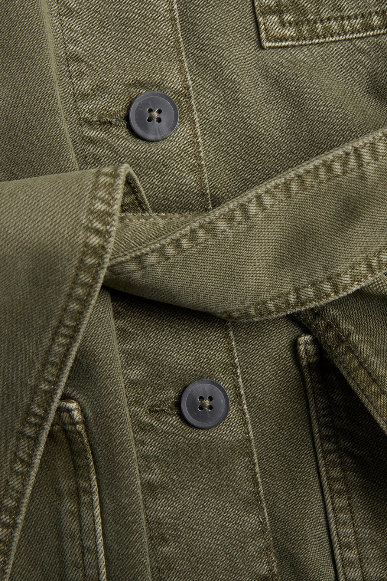 Lähikuva vihreästä twill-utilitytakista, jossa sidottu kangasvyö ja tummat napit.