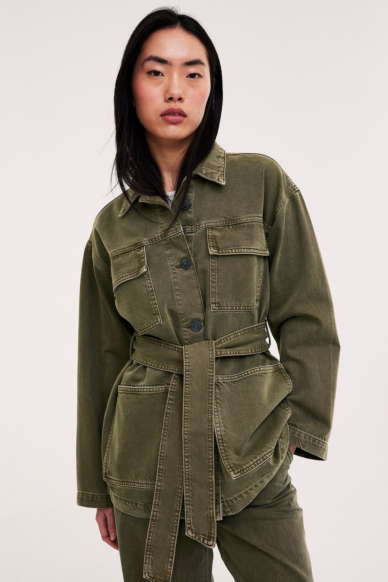 Widok z przodu: Kobieta w zielonej bawełnianej kurtce z kieszeniami i paskiem, plus zielone spodnie.