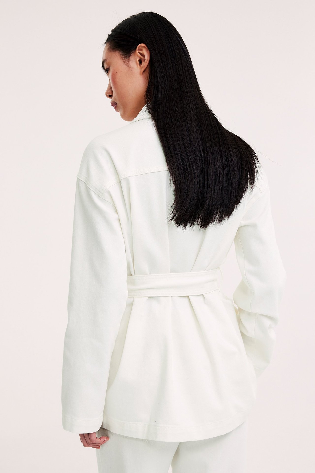 Naisen takanäkymä offwhite twill-utilitytakissa, jossa solmittu vyö ja yhteensopivat housut.