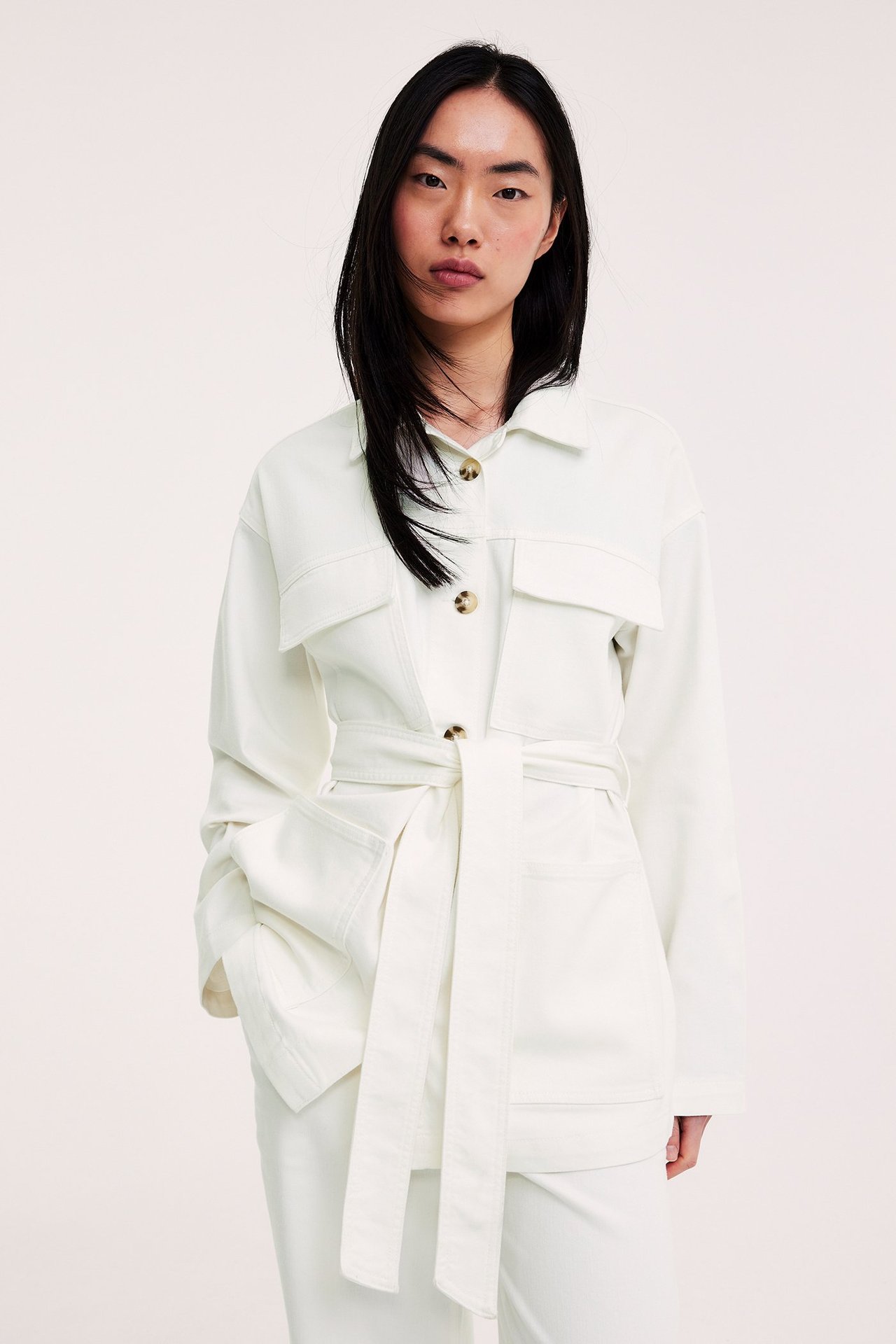 Naisen etunäkymä päällä luonnonvalkoinen utilitytakki läppätaskuilla ja solmitulla vyöllä.