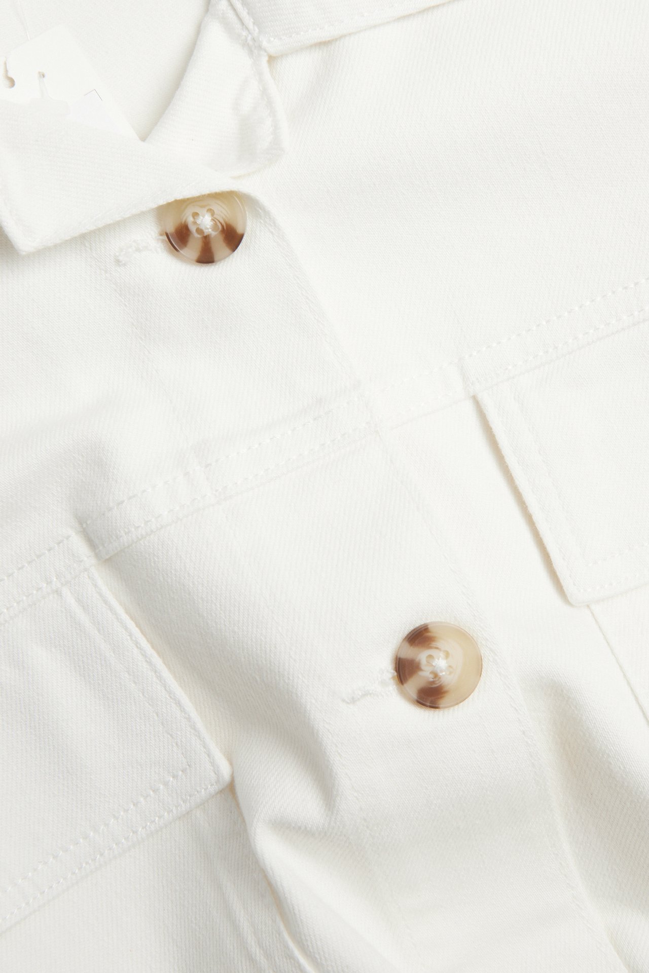 Närbild av en offwhite twill utilityjacka med bruna knappar och påsydda fickor.