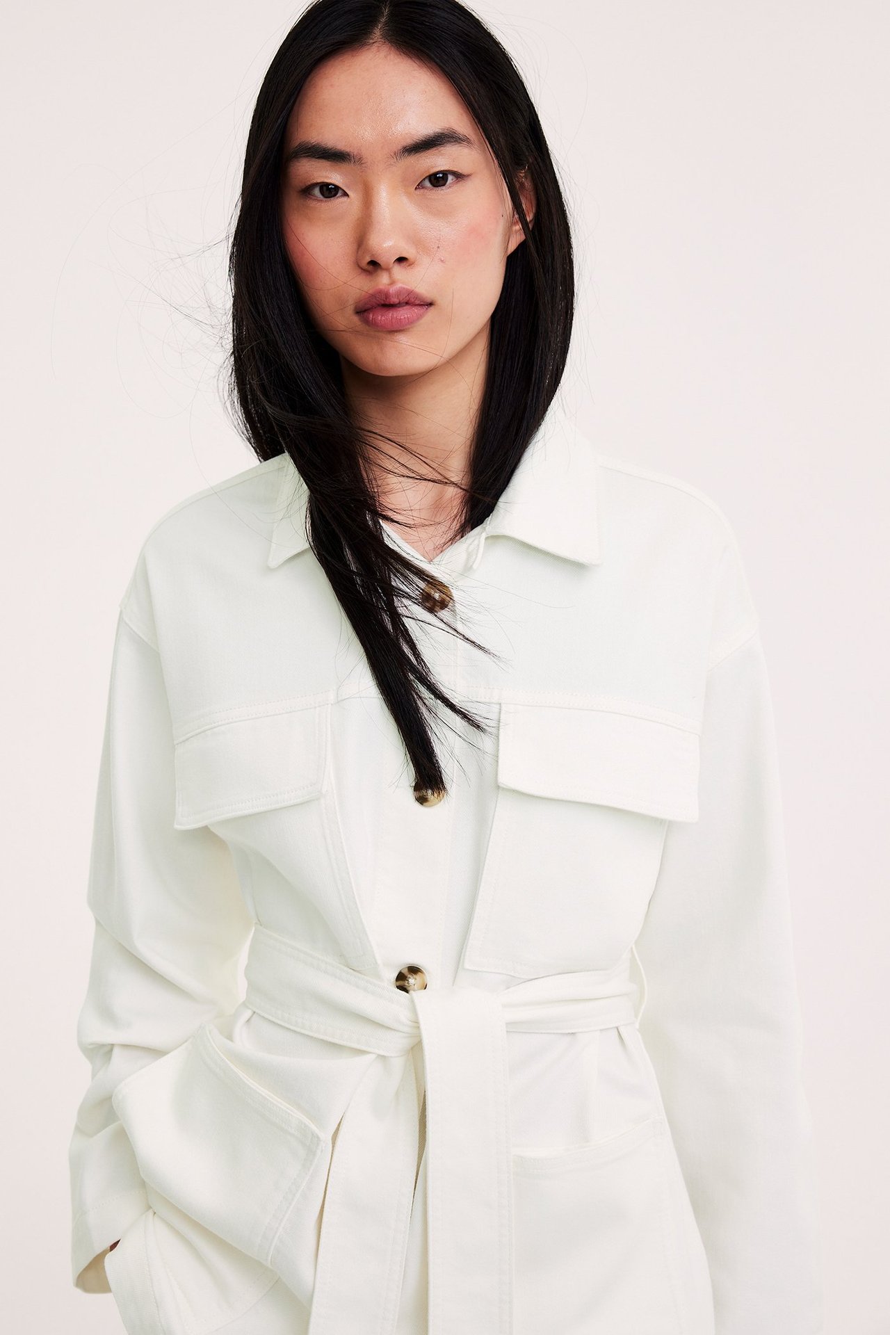 Frontvisning av en kvinne i en offwhite twill-utilityjakke med knytebelte og synlige knapper.